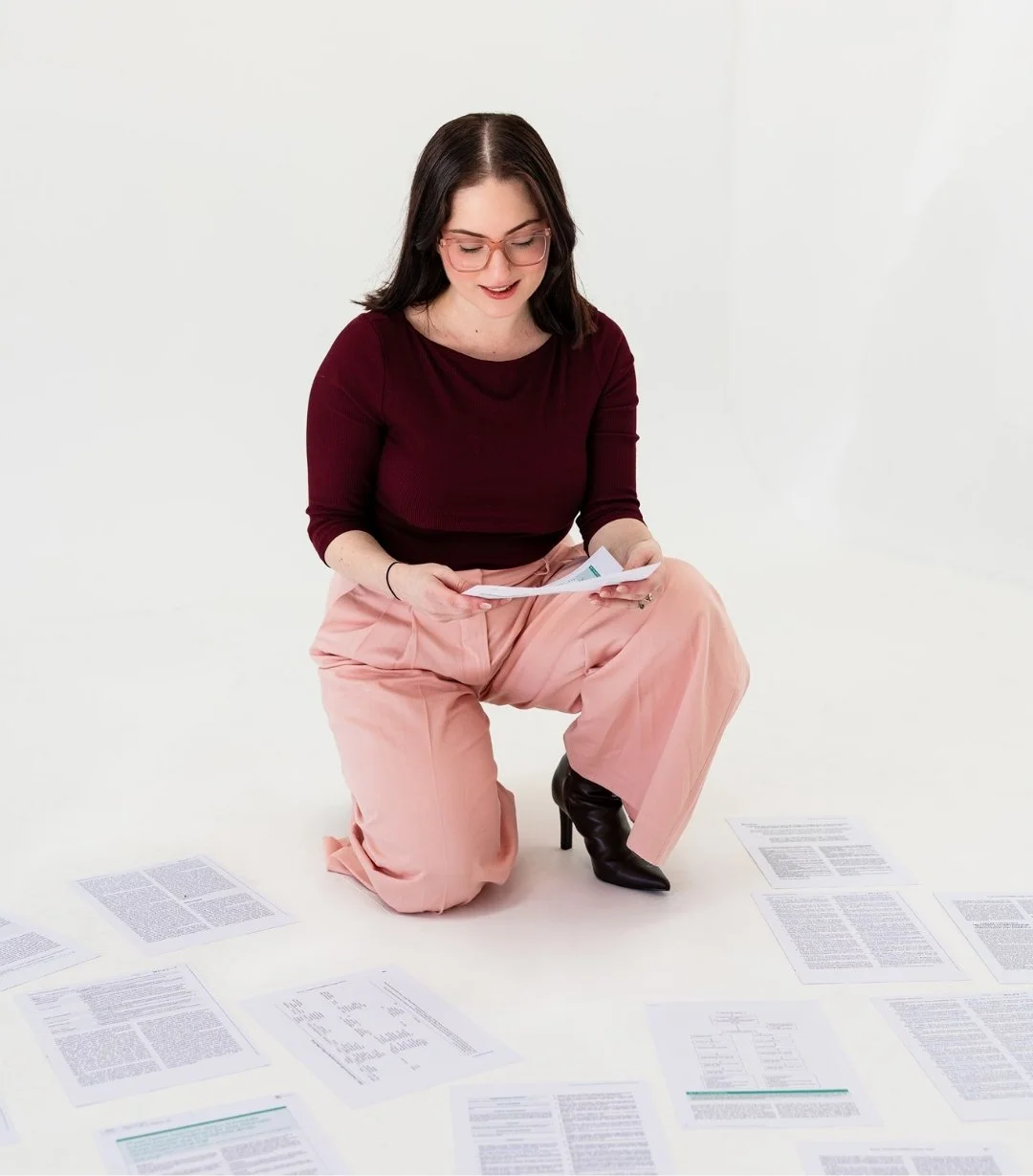 Woman wearing glasses, a maroon top, and pink pants, squatting on the floor, looking at papers spread out around her.