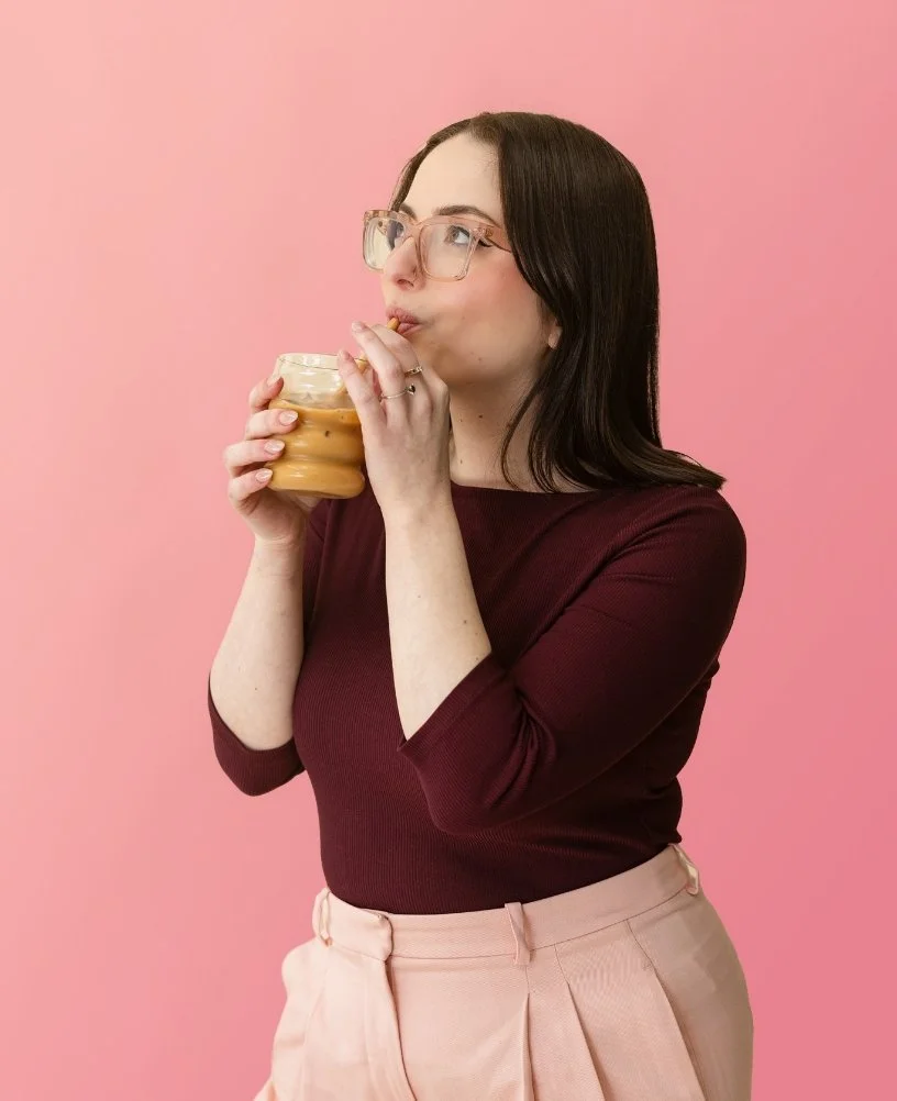 Woman with glasses drinking iced coffee from a glass jar against a pink background.