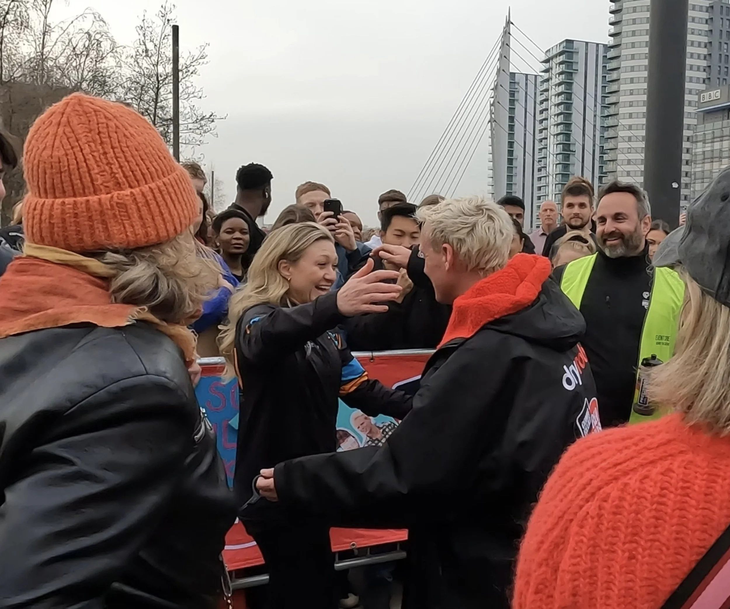A diverse group of people gathered outdoors, some smiling and interacting, with modern buildings and an overcast sky in the background. One person is wearing a bright orange knit hat and a dark jacket, while another has gray hair and a high-visibility vest.