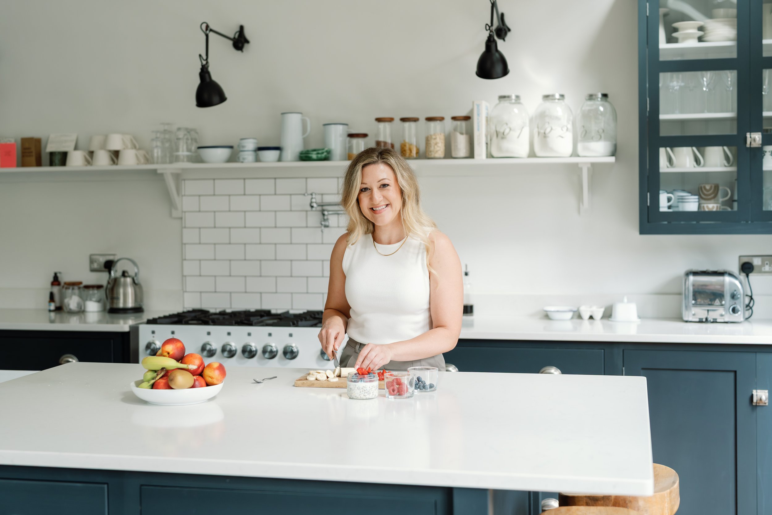 A woman with blonde hair, wearing a white sleeveless top, smiling in a modern kitchen. She has sliced bananas and strawberries on a cutting board in front of her. There are bowls of berries, a bowl of apples, and bananas on the white kitchen island. 