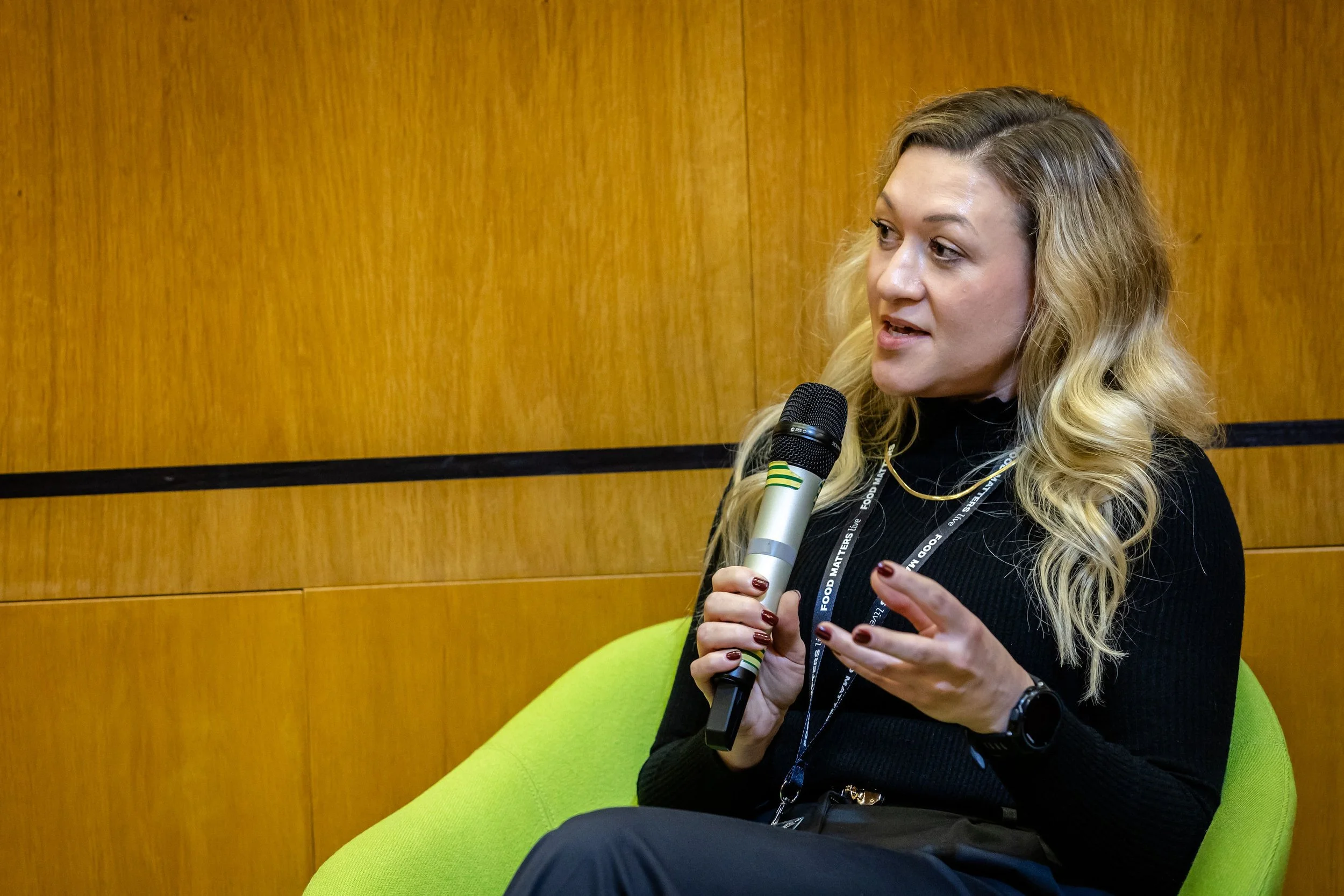 Woman with long wavy hair holding a microphone and speaking in front of a wooden wall, sitting on a green chair.