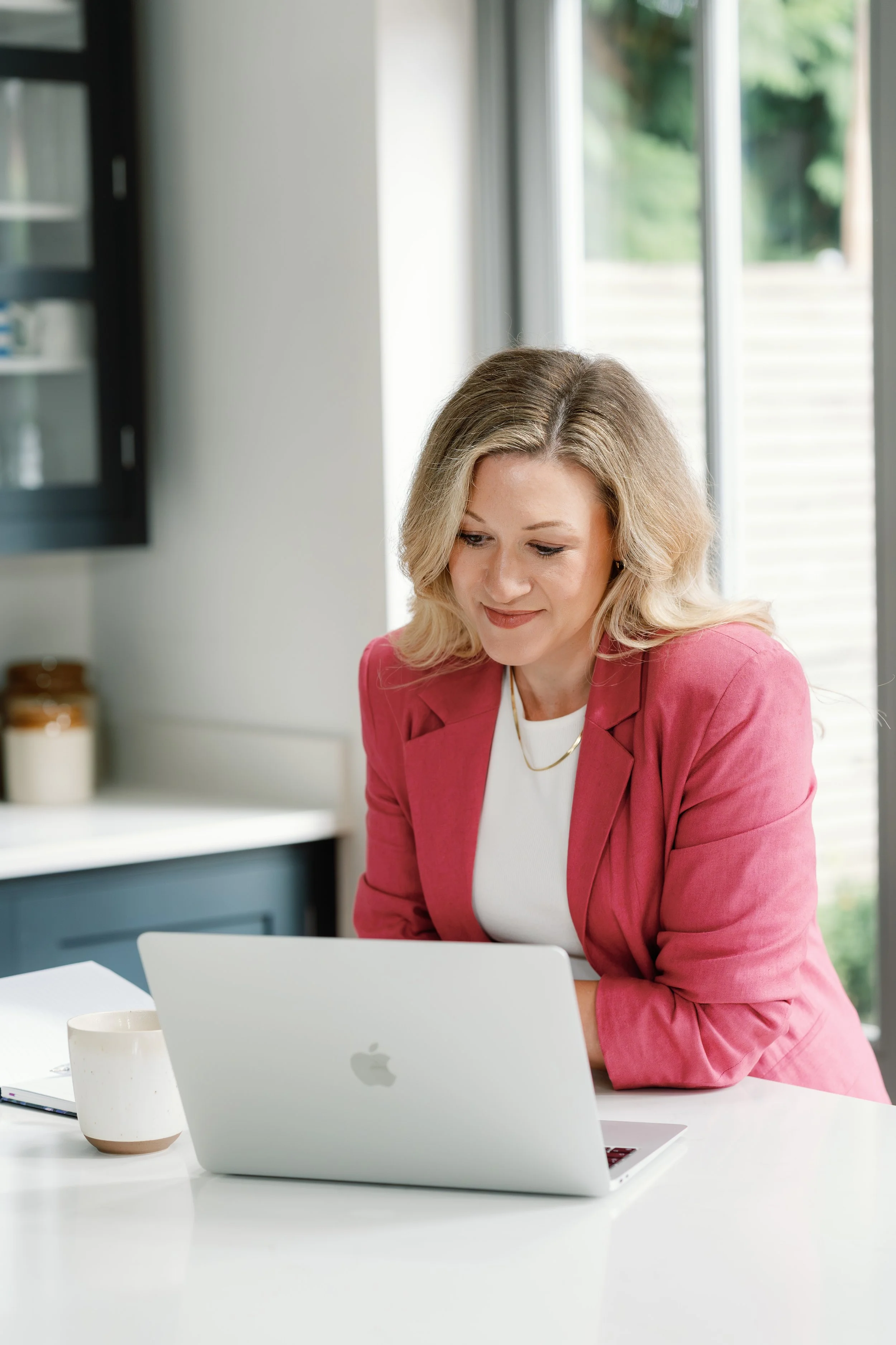 A woman with blonde hair wearing a pink blazer working on a silver laptop at a white desk in a bright room with large windows.