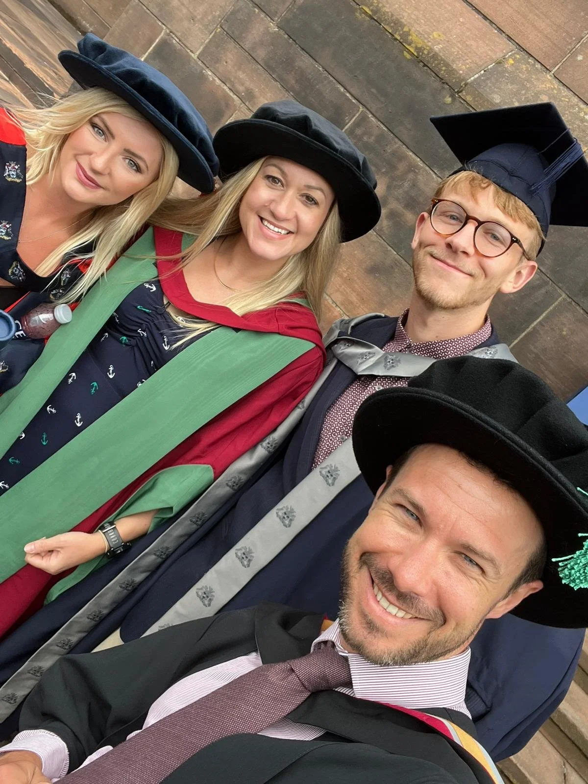 Four young adults in graduation caps and gowns taking a selfie outside against a brick wall, celebrating their graduation.
