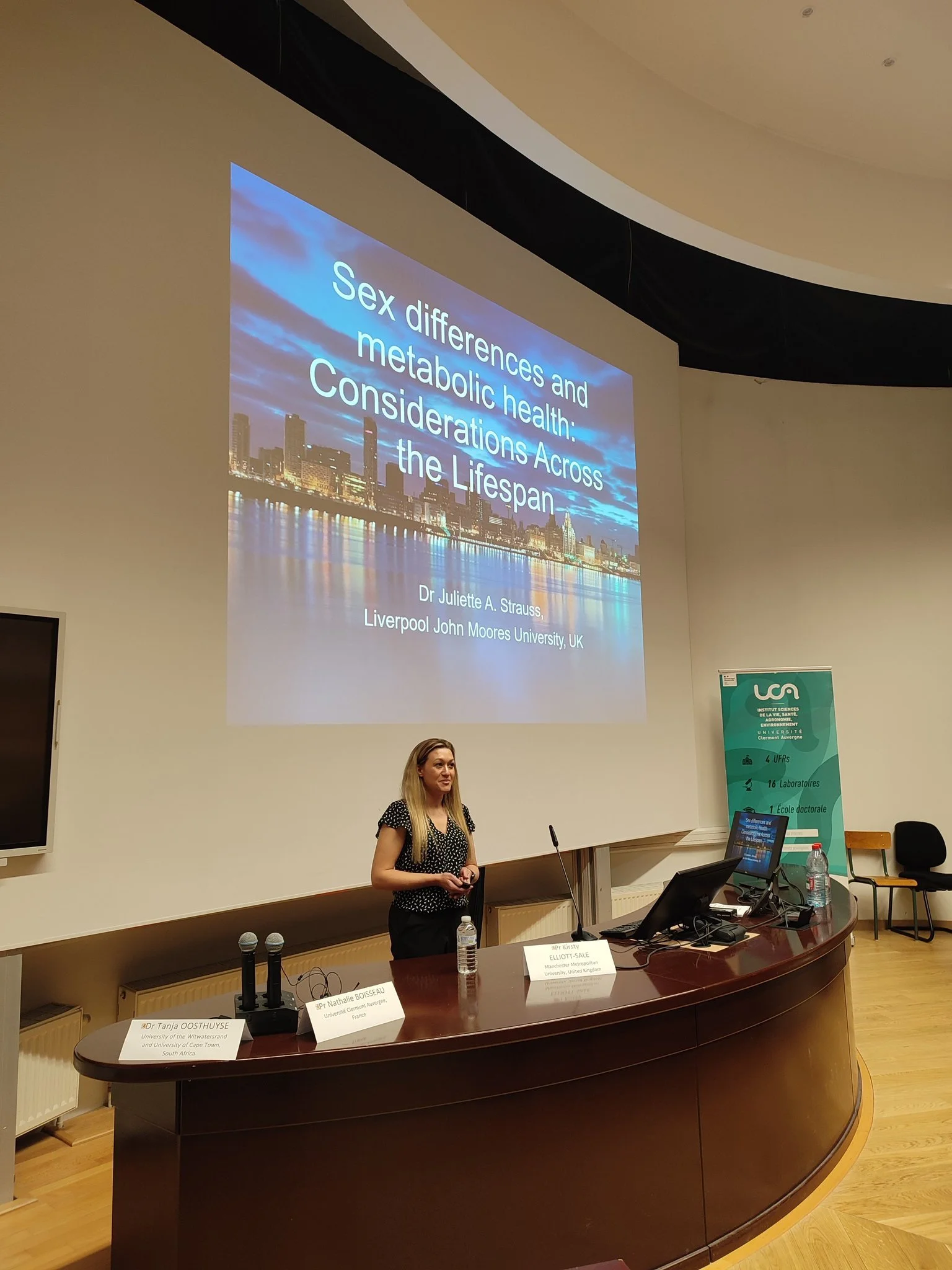 A woman is giving a presentation in a conference room, standing in front of a large screen displaying a slide titled 'Sex differences and metabolic health: Considerations Across the Lifespan.' There are desks with name cards, microphones, and laptops, and a banner on the side wall indicating an academic event.