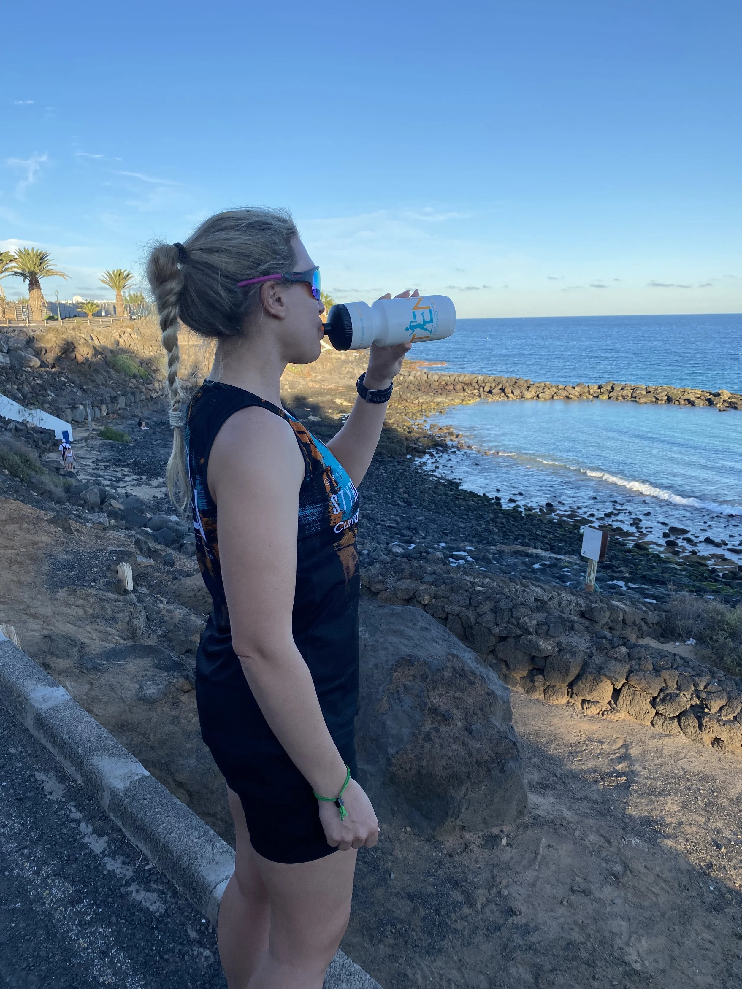A young woman with blonde hair in a braid, wearing sunglasses and a sleeveless athletic top, drinks water from a white water bottle while standing on a rocky coastal path with the ocean in the background.