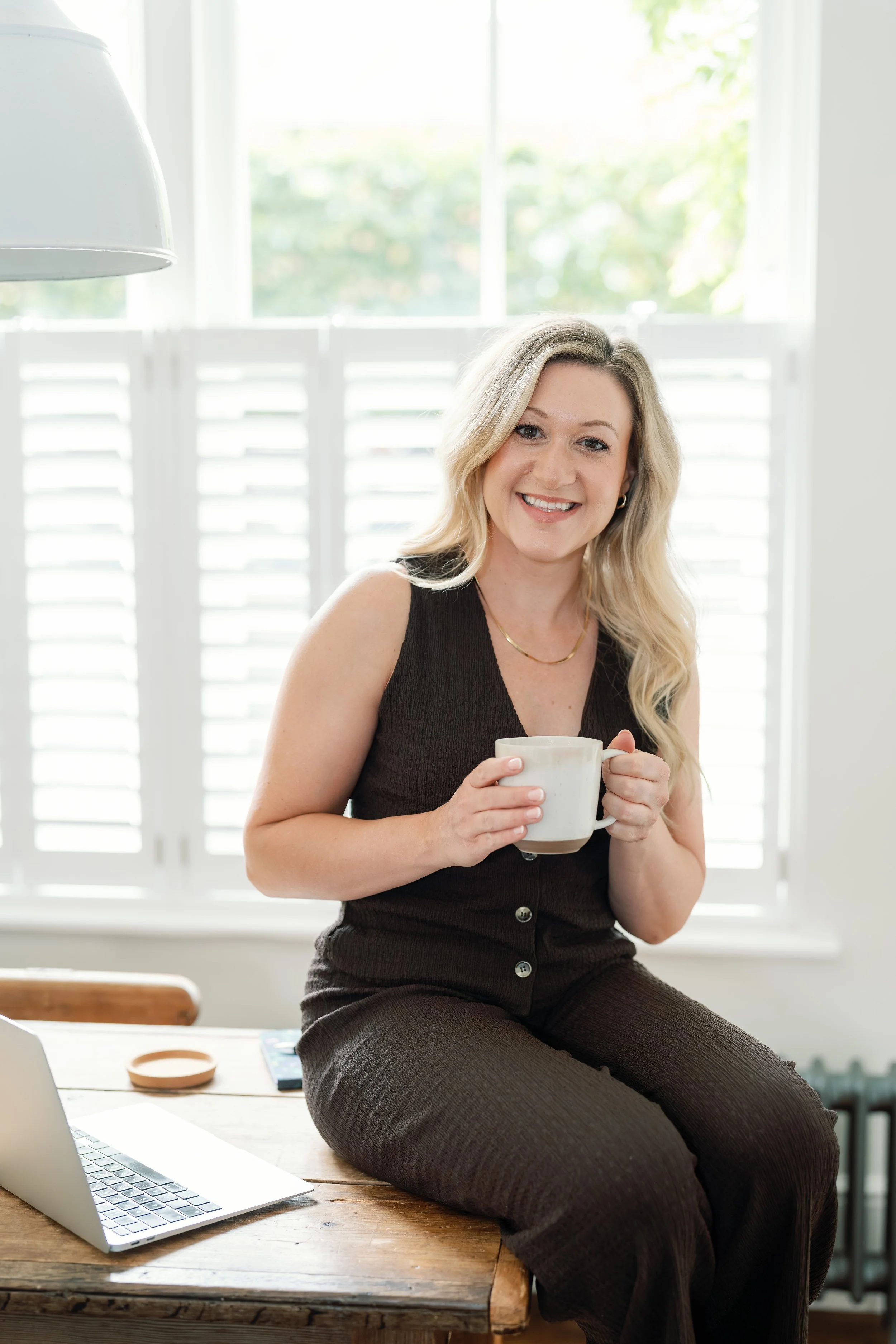 A woman with blonde hair sitting on a wooden table, holding a white mug, smiling at the camera, with a laptop on the table and a window with shutters in the background.