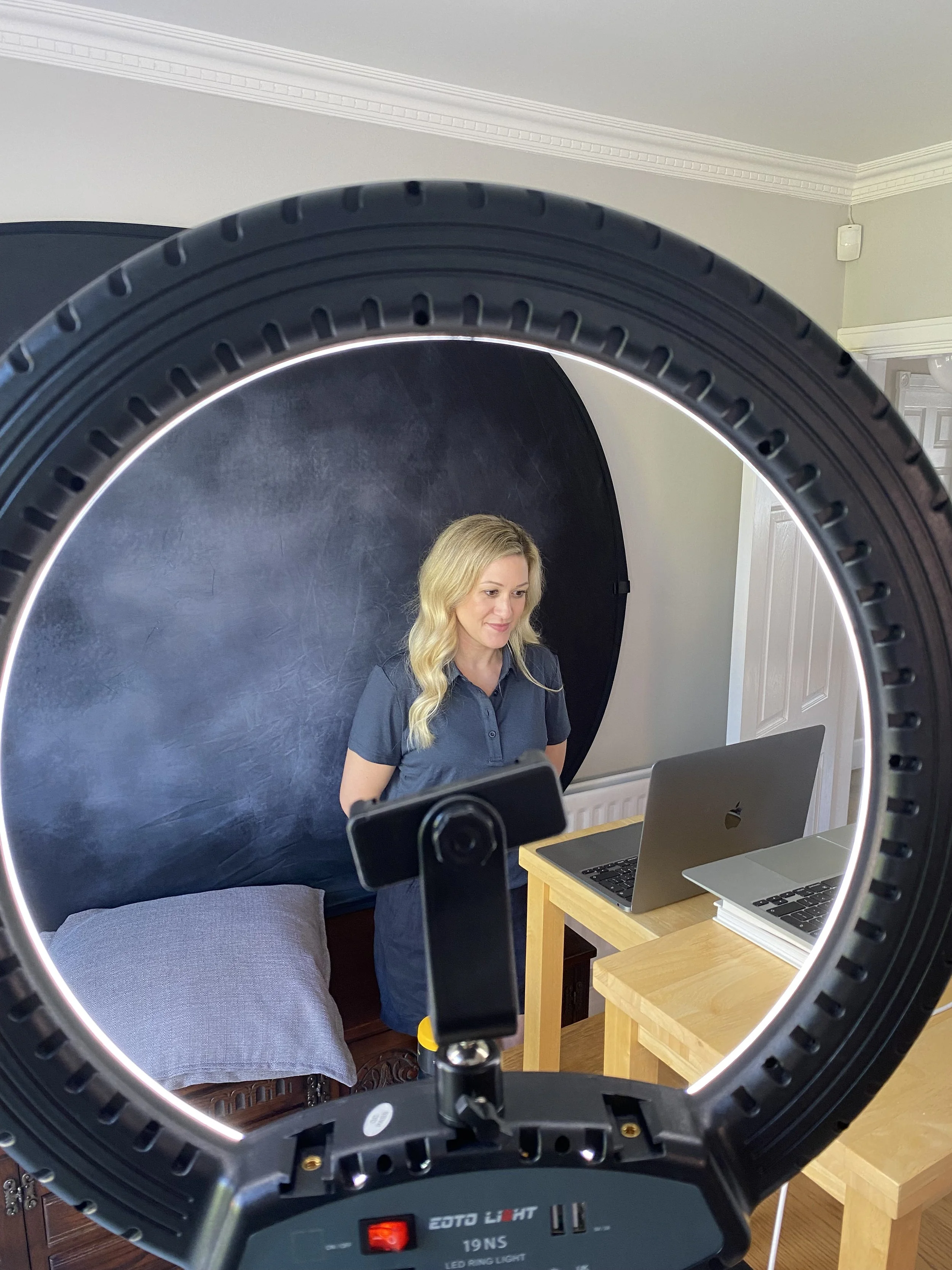 A woman with blonde hair working on a laptop, framed by a circular ring light, with a black backdrop behind her in a room with white crown molding.