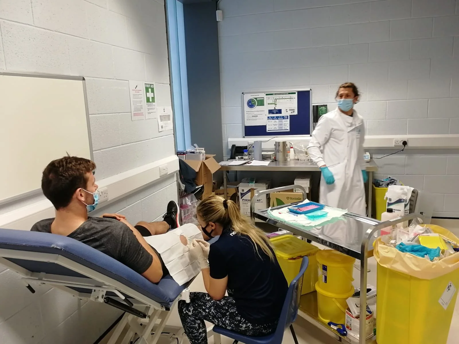 A person is lying on a hospital bed with their arm extended, receiving a medical procedure, while a medical professional checks the patient. Two other people are seated at the bedside, wearing face masks. Medical supplies and equipment are visible in