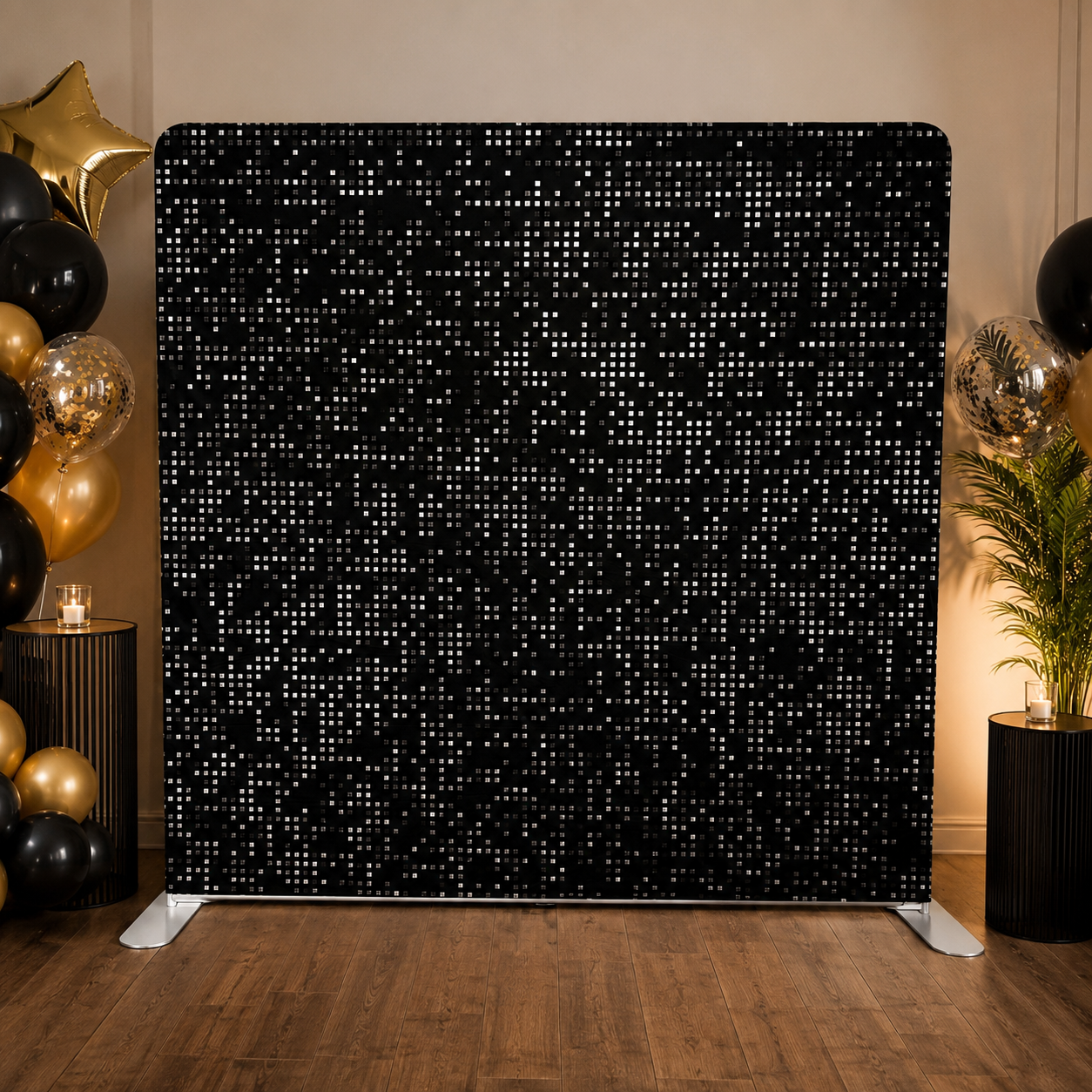 Decorative party backdrop with black and white pixel pattern, surrounded by gold and black balloons, candles, and a potted plant.