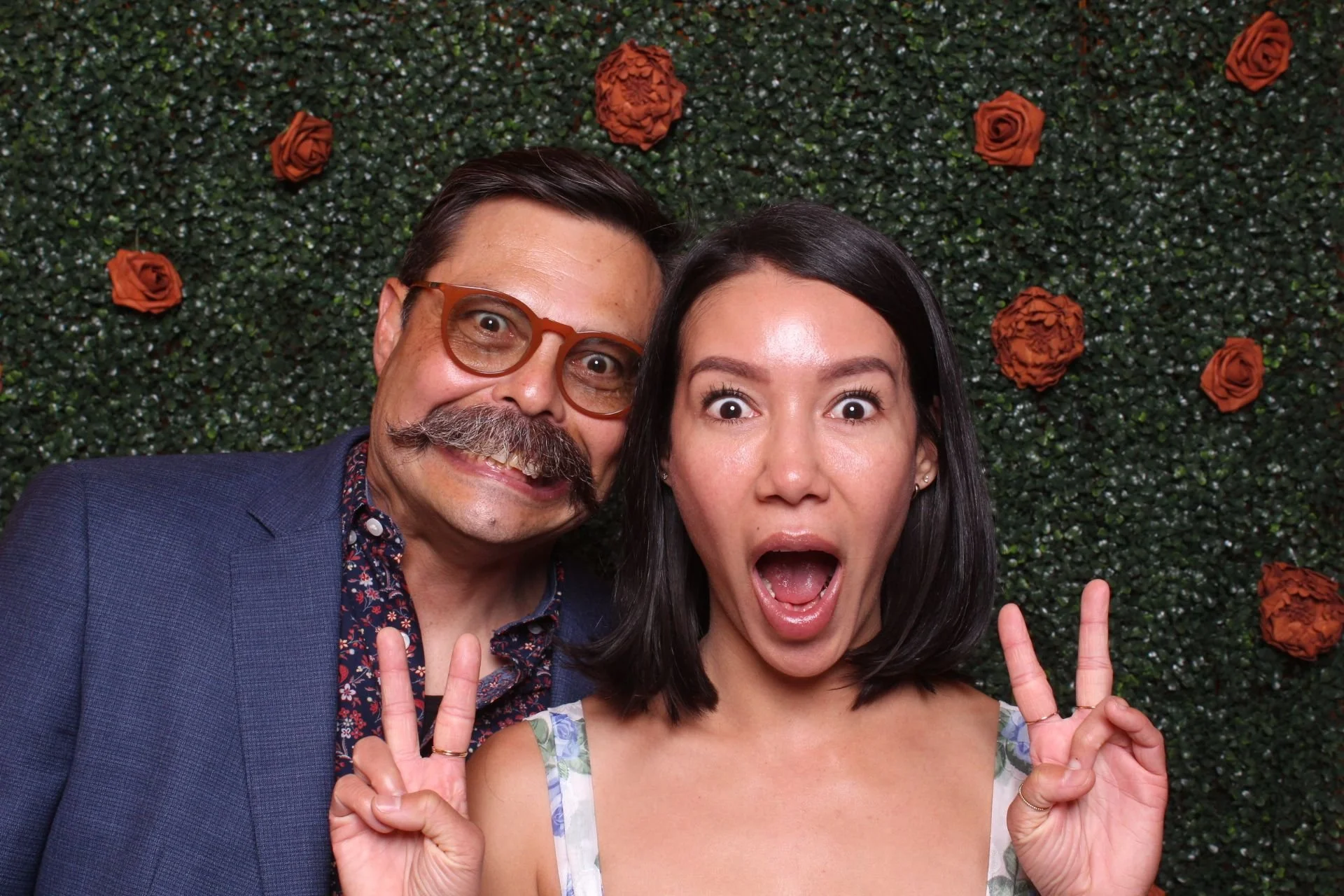 A man with glasses, a mustache, and a beard making a surprised face next to a woman with dark hair taking a selfie in front of a green hedge decorated with orange roses, both showing peace signs.