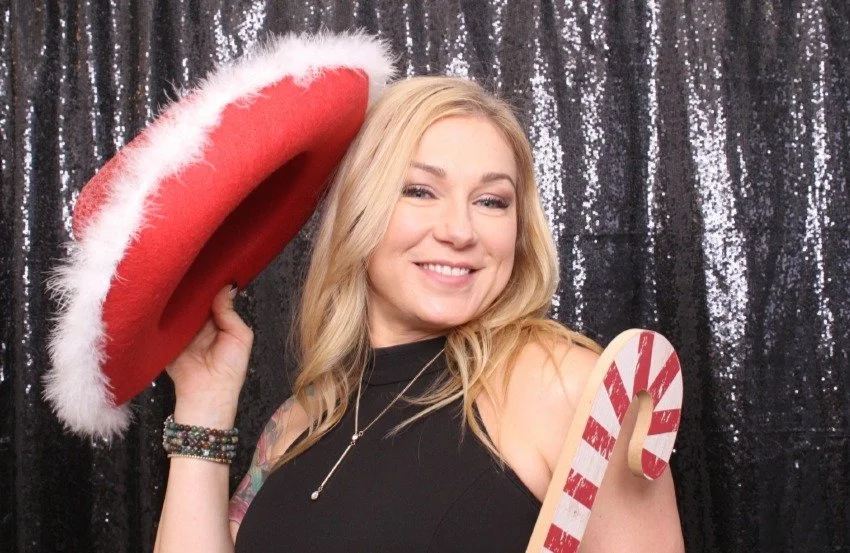 A woman with blonde hair smiling, wearing a black top, holding a red Santa hat with white trim in one hand and a candy cane in the other, standing in front of a black, shiny, sequin background.