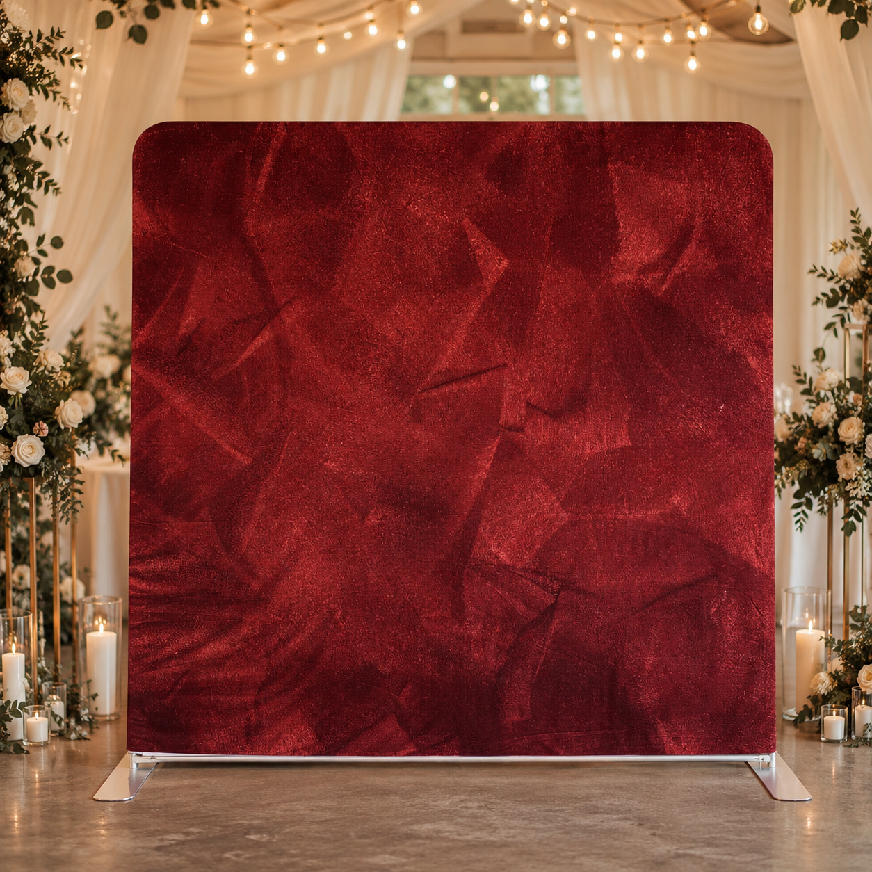 A large red velvet photo backdrop with a textured surface, set up at a decorated event venue with floral arrangements and lit candles surrounding it.
