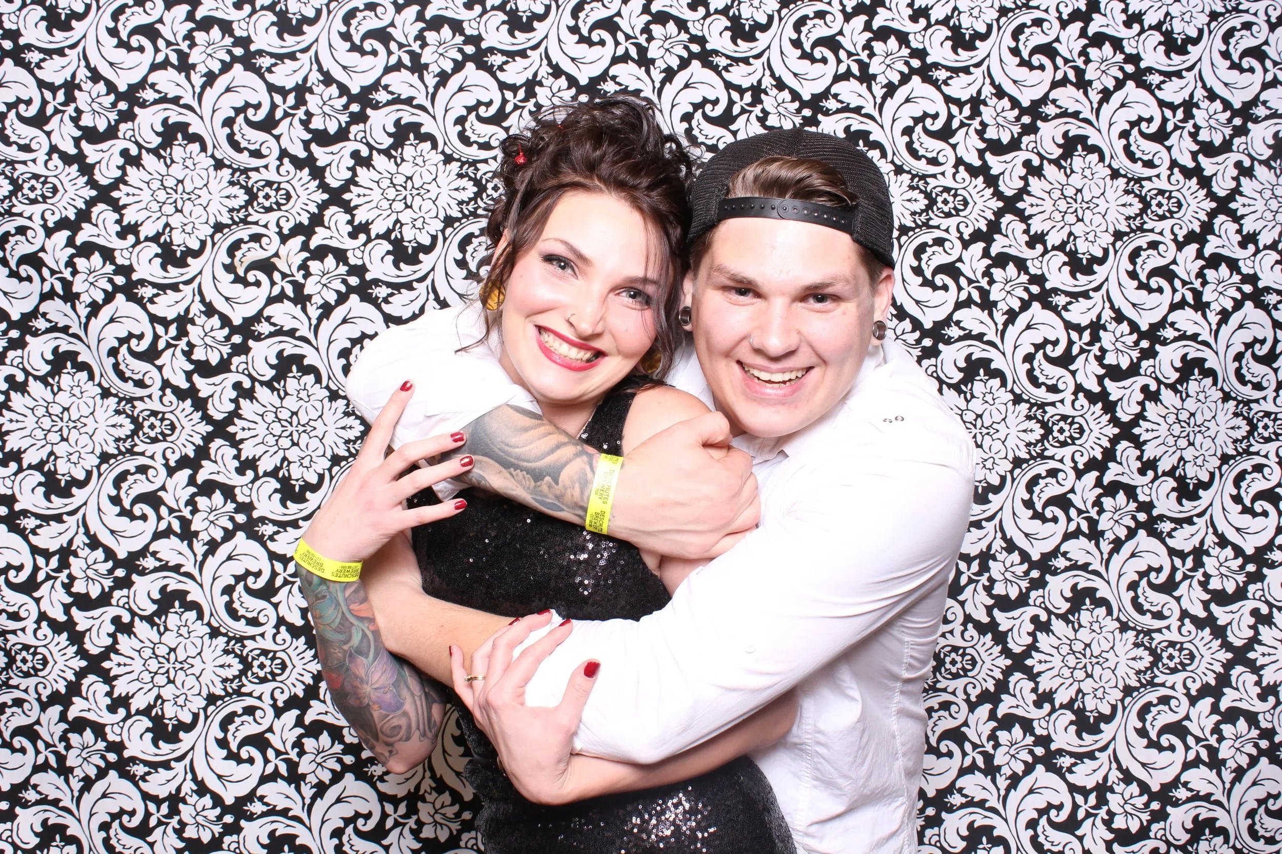 Two smiling people hugging in front of a black and white floral patterned wall. The woman on the left has dark, wavy hair, red lipstick, and tattoos on her arm. The man on the right is wearing a black cap backward, earrings, and a white shirt.