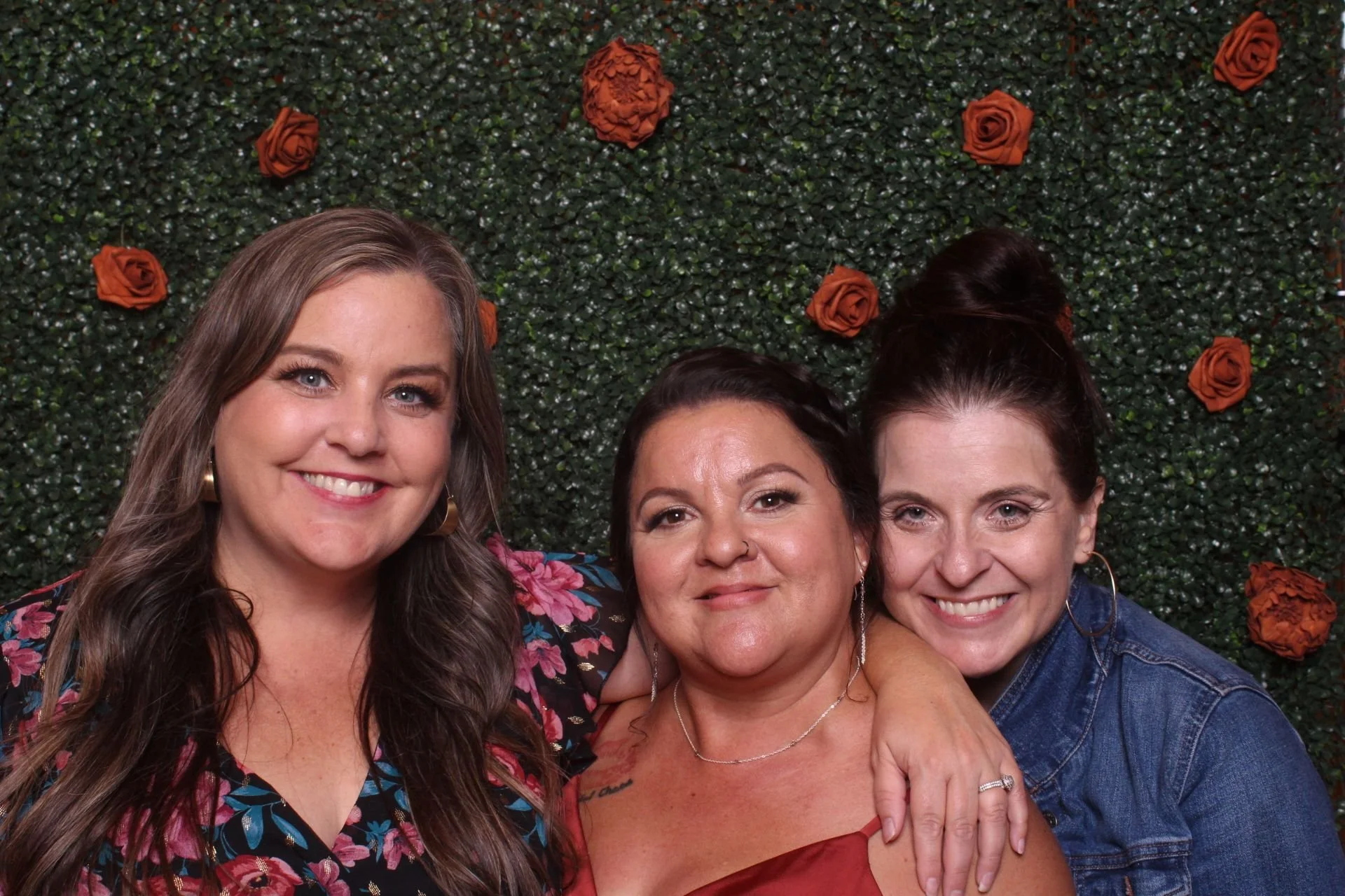 Three women smiling, standing close together in front of a green hedge with orange roses, with their heads and shoulders visible.