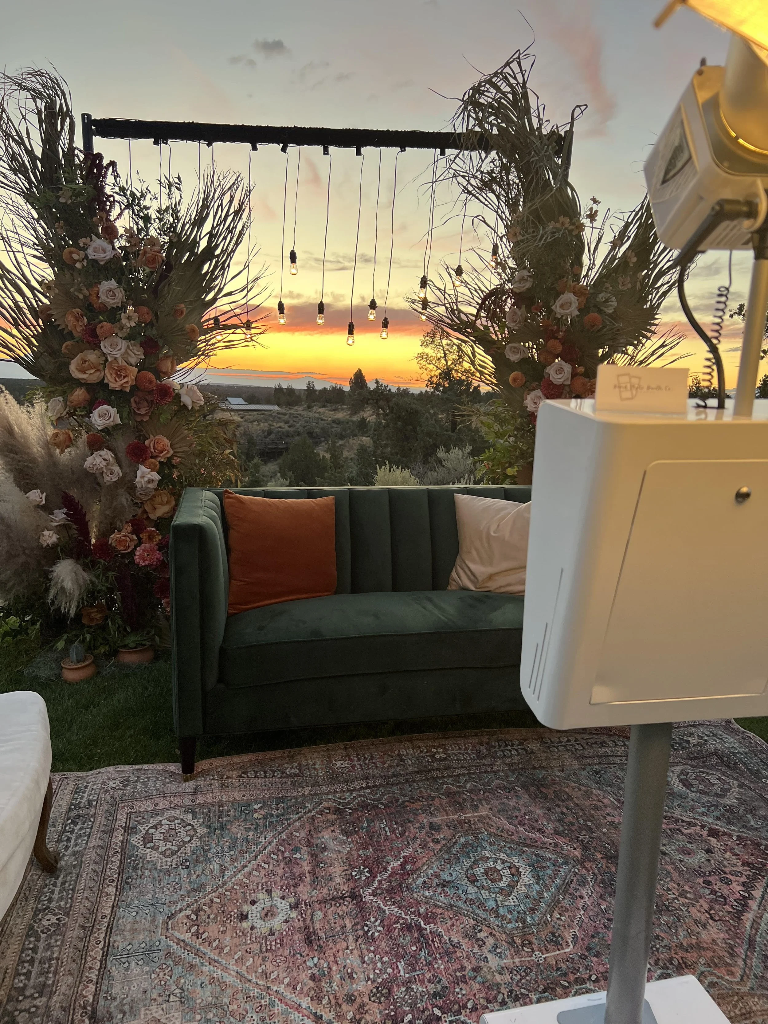 An outdoor setup at sunset with decorative floral arrangements on either side and hanging string lights, featuring a green sofa with orange and cream pillows, an ornate rug, and a view of the sunset over a landscape with trees and hills.