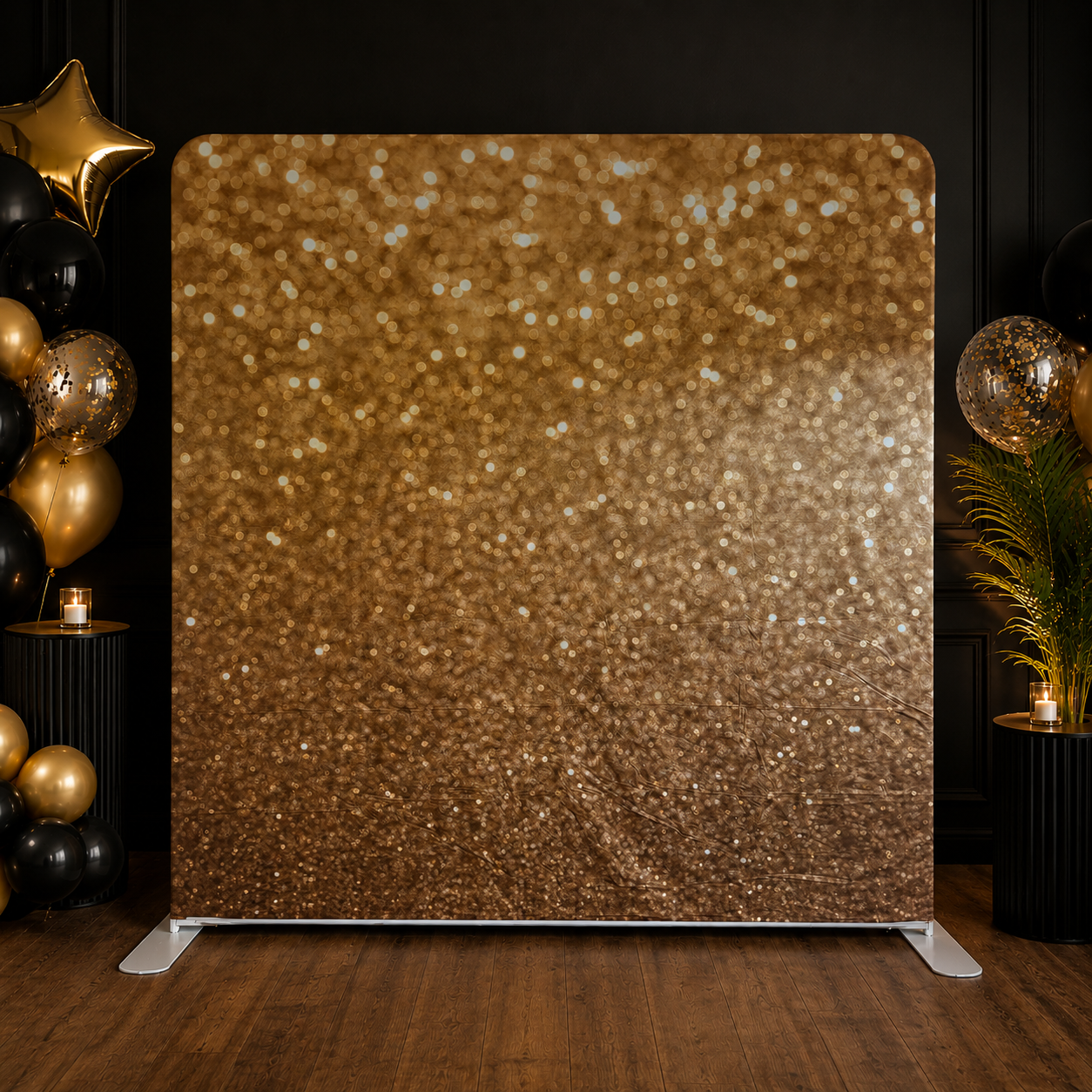 A gold glittered backdrop with black and gold balloons beside it, decorated with small candles and green plants on black tables.