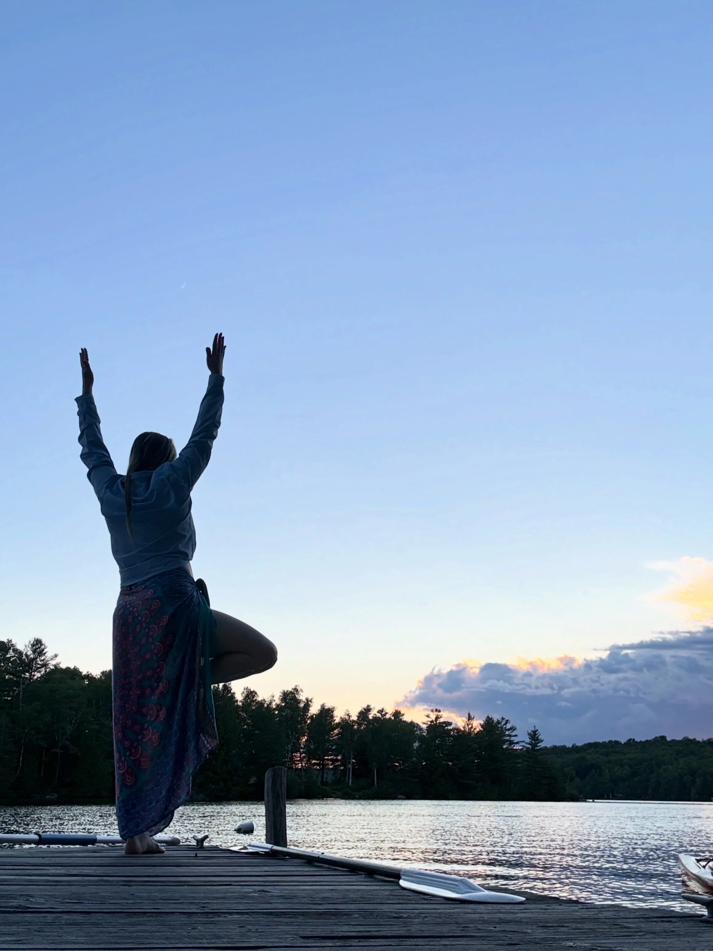 A person practicing yoga on a dock by a lake during sunset, standing on one leg with arms raised, wearing a long skirt, with trees and clouds in the background.