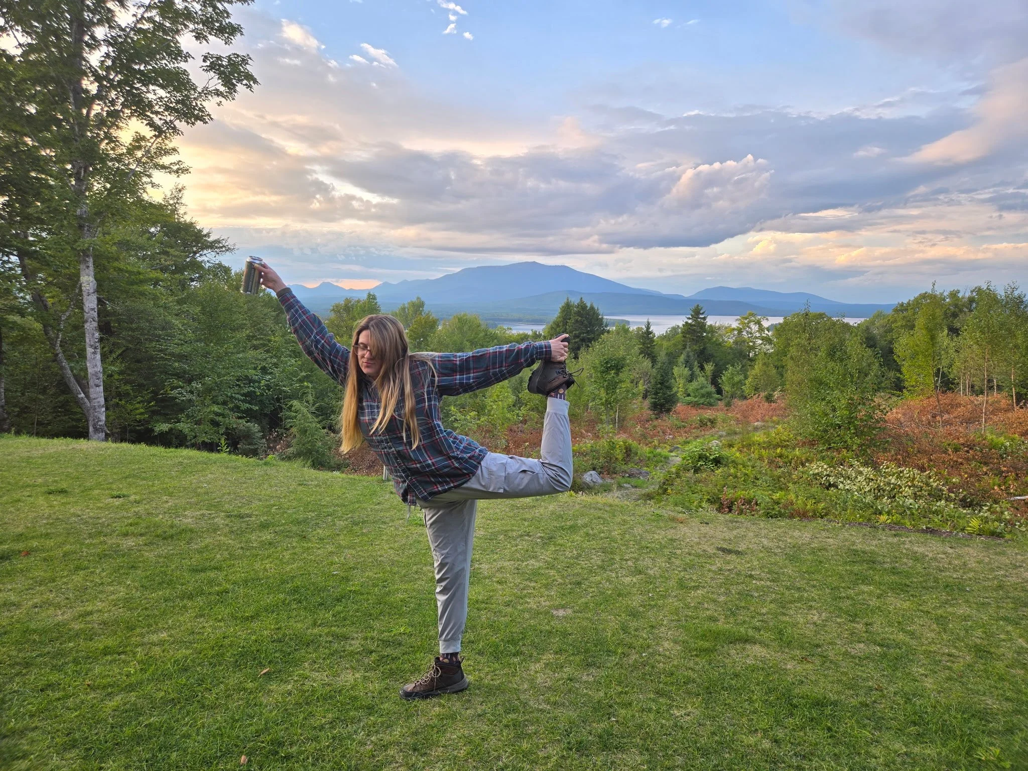 A woman practicing yoga outdoors in a park with green grass, trees, and mountains in the background, performing a standing bow pose while holding a beverage can in one hand.