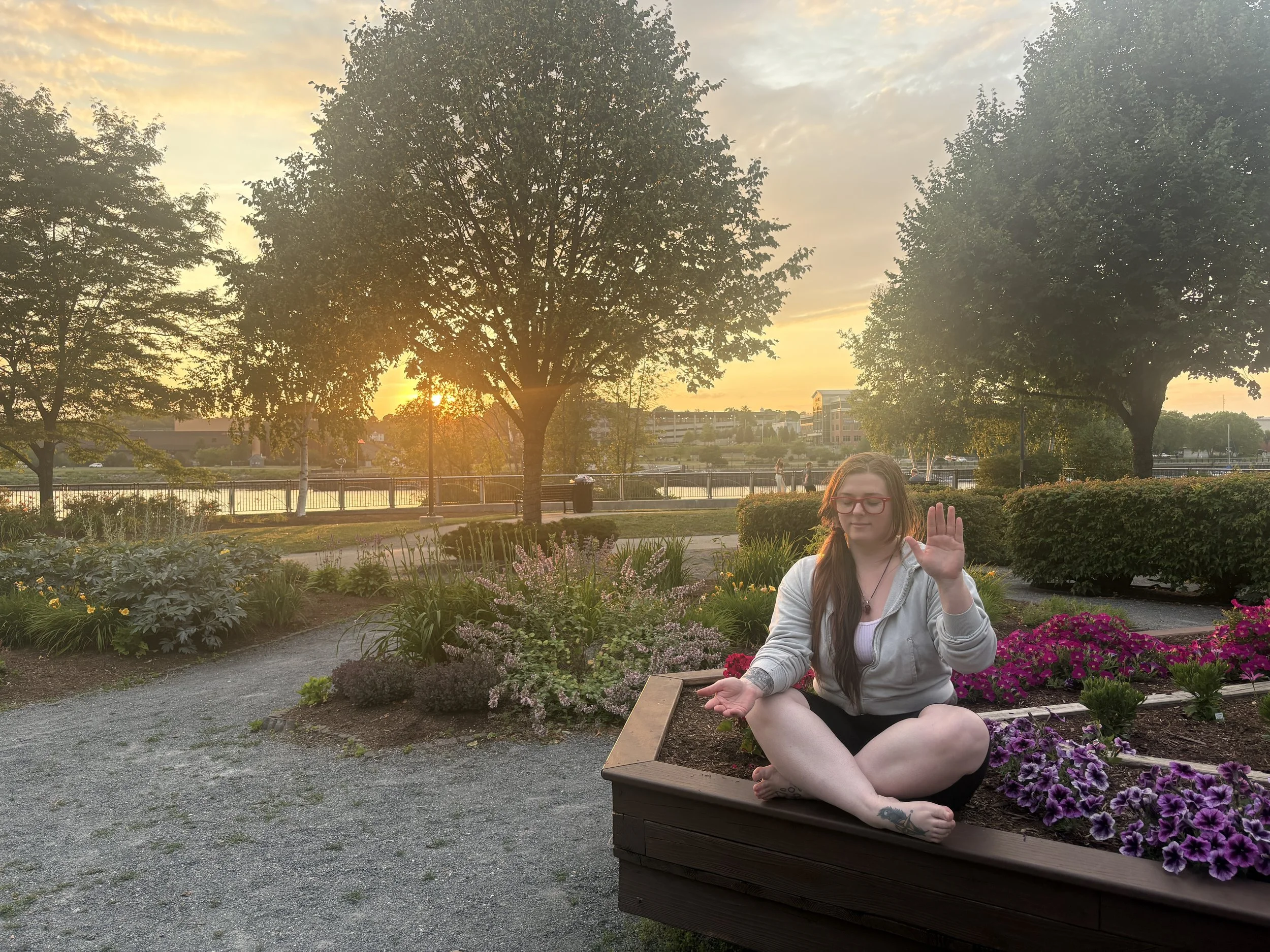 A young woman with long dark hair and glasses is sitting cross-legged in a flower bed with one hand up and one hand down, palms open. The scene is outdoors during sunset, with trees, a garden, and a park in the background.