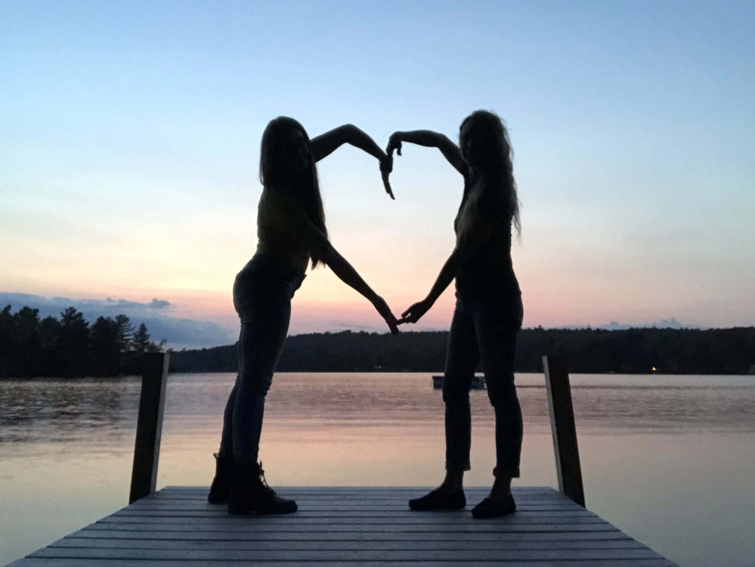 Two women holding hands and forming a heart shape with their arms on a dock over water at sunset or dusk