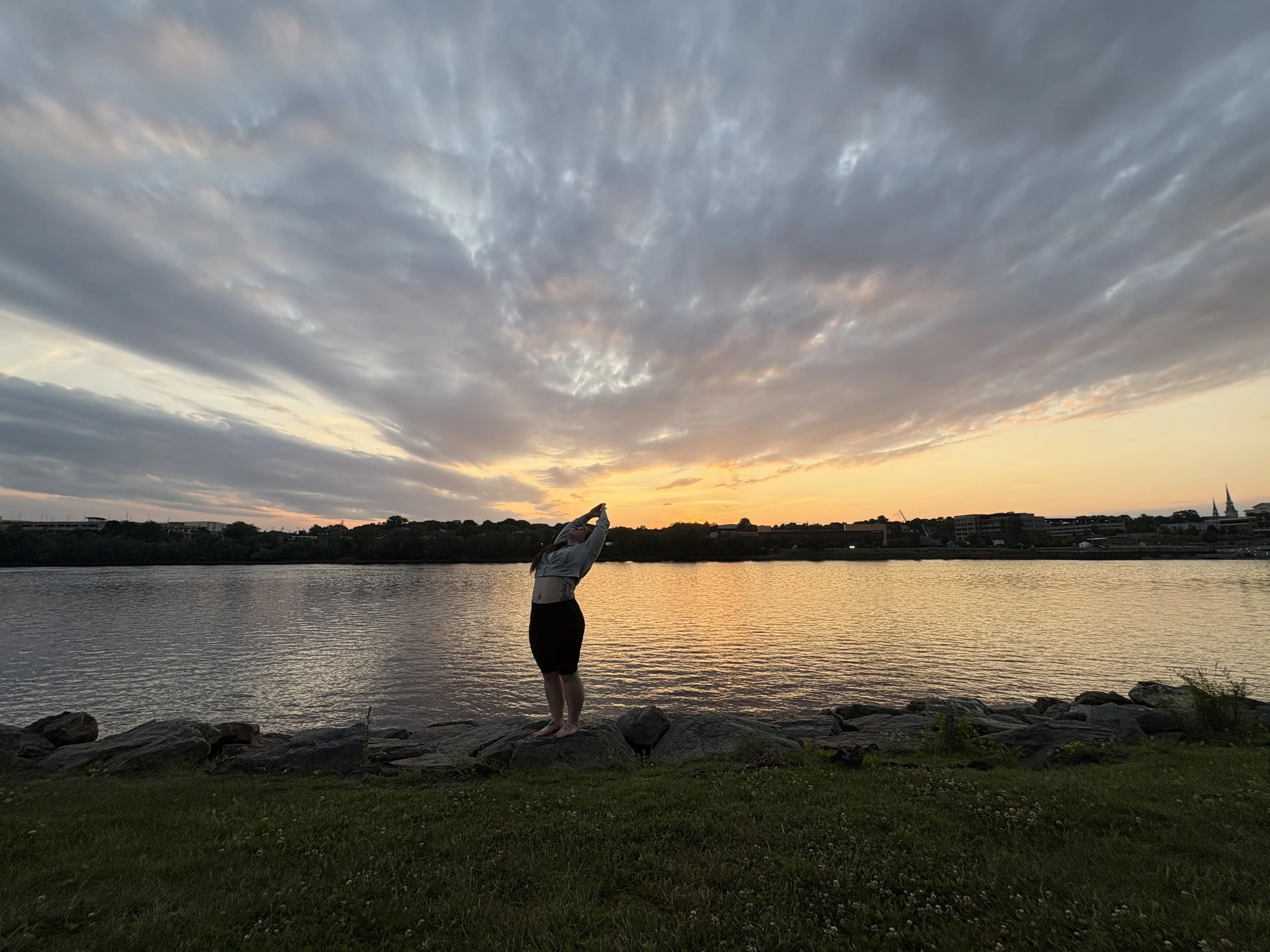 A person standing on rocks by the water during sunset, doing yoga as the sky is filled with clouds and the sun is setting behind the horizon.