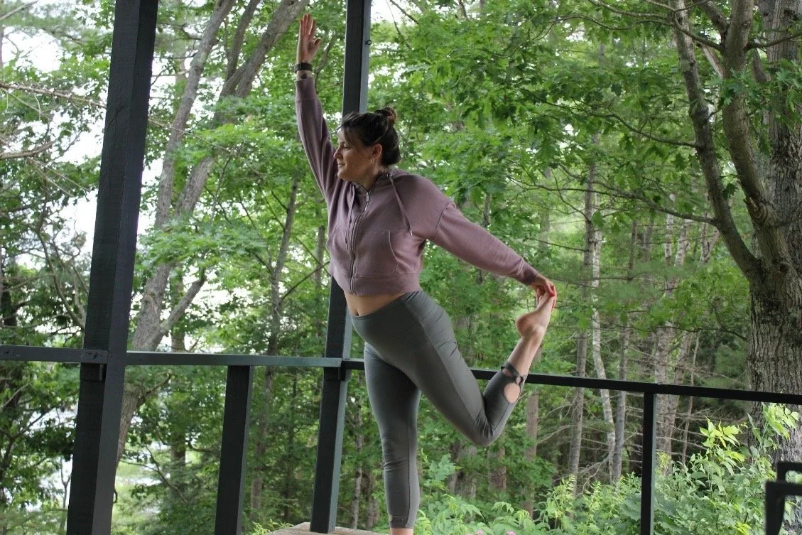 A woman practicing yoga outdoors on a balcony surrounded by trees, doing a standing bow pose while holding her foot behind her back.