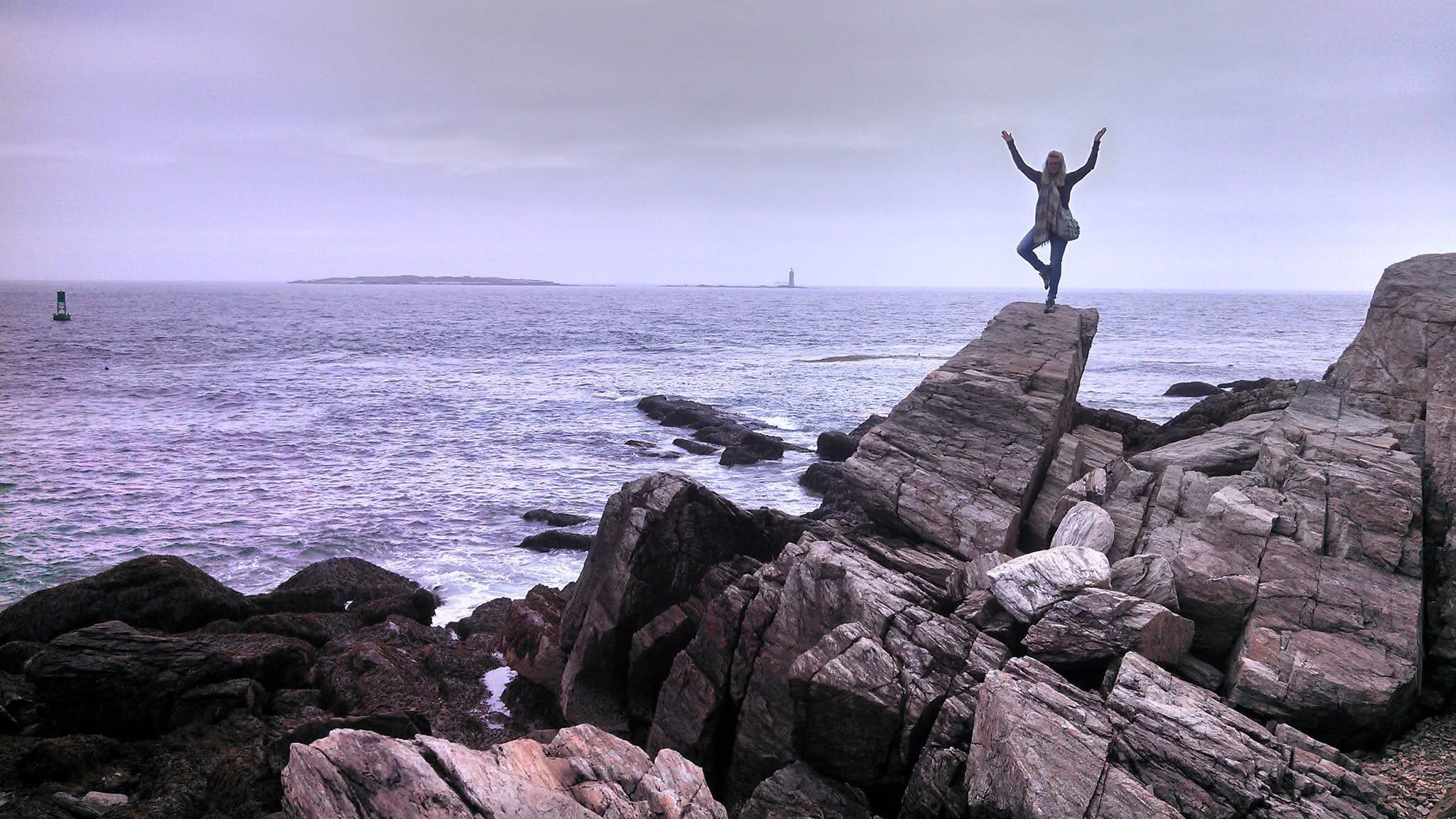 A woman standing on a large rock formation at the edge of the ocean with her arms raised. The sky is cloudy, and a lighthouse is visible in the distance across the water.