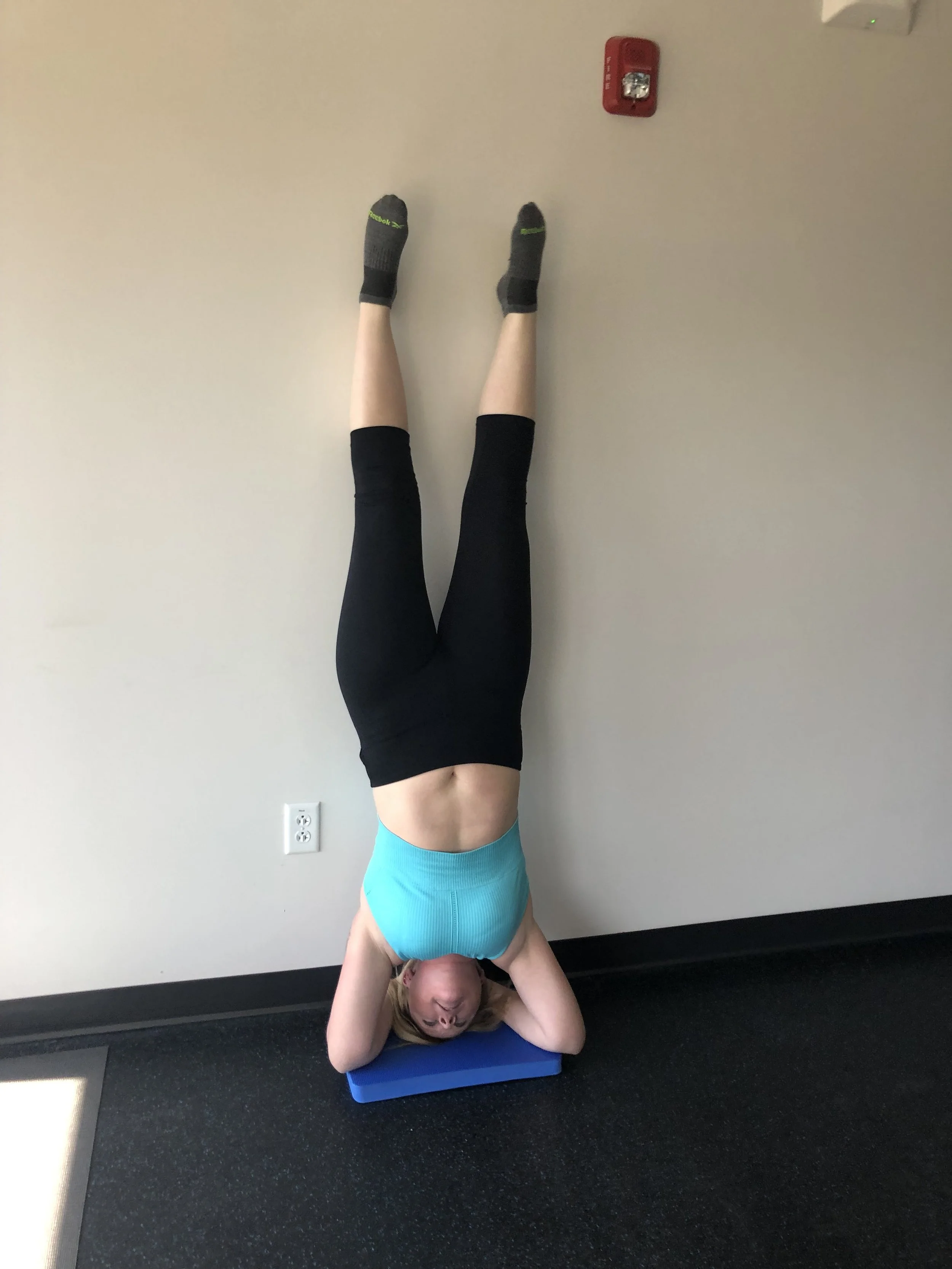 A woman doing a headstand against a beige wall in a gym. She is supporting herself with her head and hands, wearing a light blue workout top and black leggings. A blue exercise mat is beneath her head.
