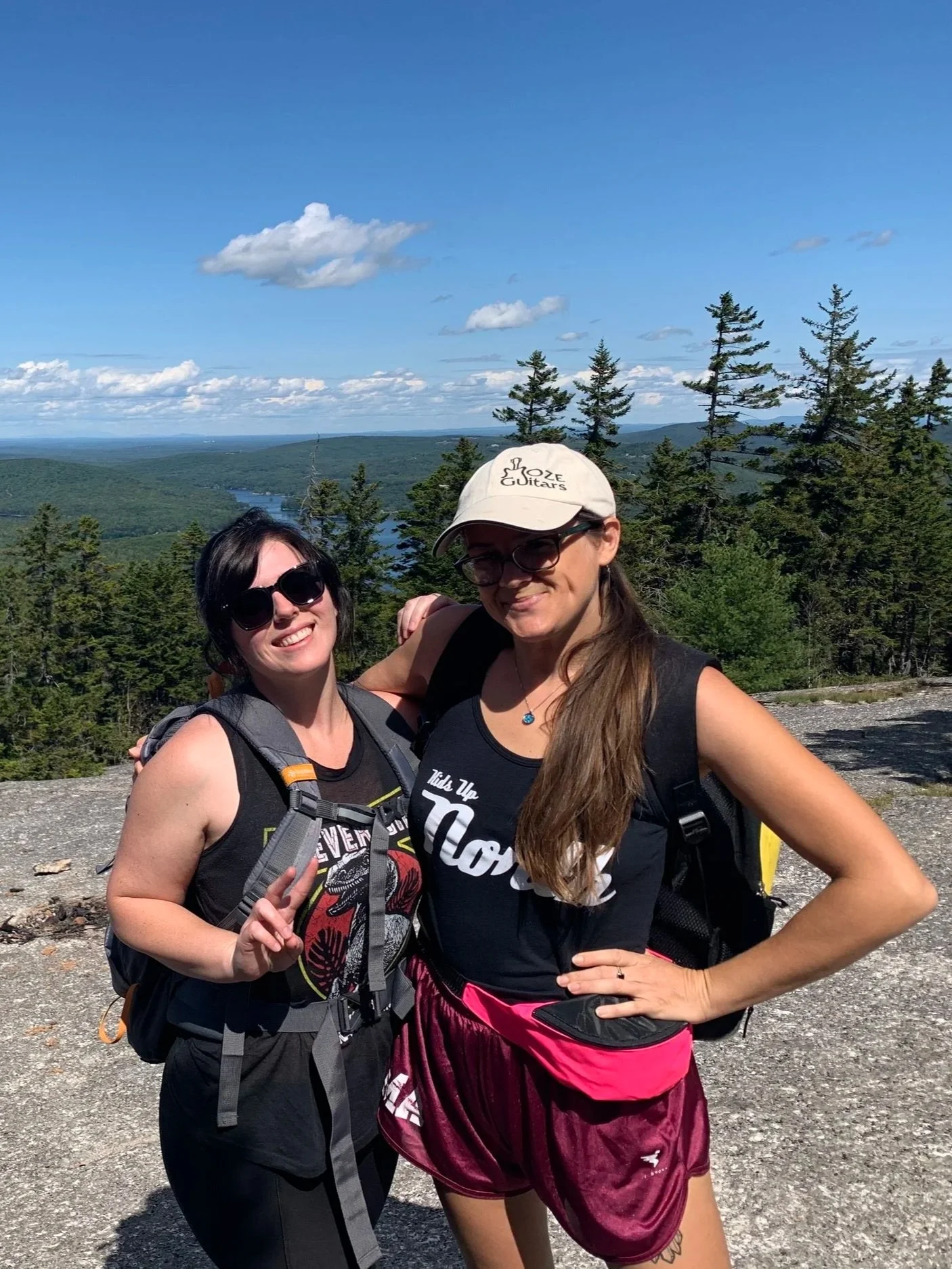Two women hiking outdoors, smiling, with a scenic view of trees, a river, hills, and a blue sky with clouds in the background.