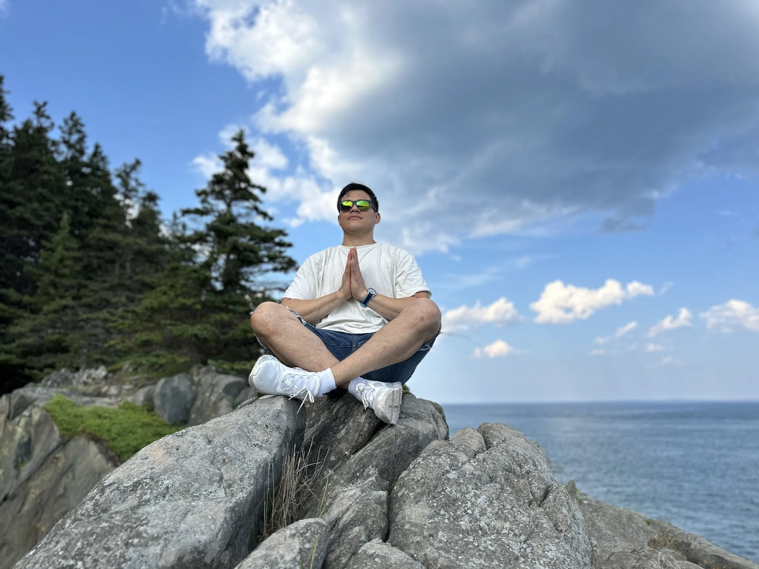 A man sitting cross-legged on rocks by the ocean, wearing sunglasses, a white t-shirt, and shorts, with hands in a prayer position, practicing meditation or yoga under a partly cloudy sky.