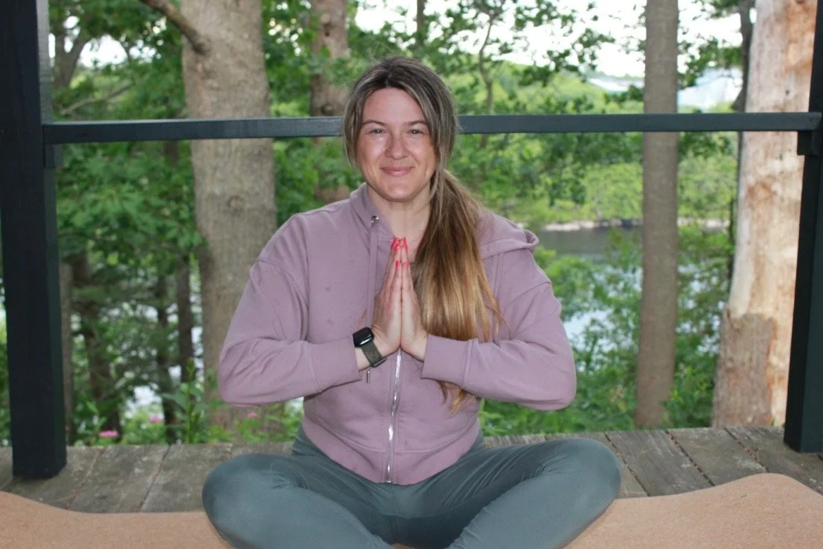 Woman practicing yoga outdoors on a wooden deck, sitting cross-legged with hands in prayer position, surrounded by trees.