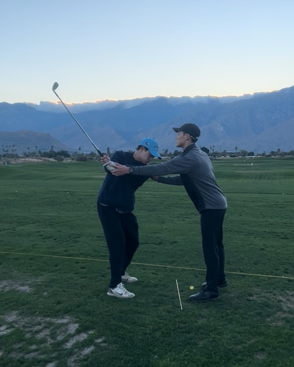 Two men on a golf course during the evening, one instructing the other on a golf swing, with mountains in the background.