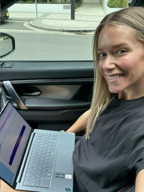 A woman with blonde hair smiling while sitting in a car with a laptop on her lap, outside on a street.