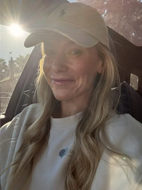 Woman sitting in a vehicle wearing a baseball cap and white shirt, with sunlight shining through the window.
