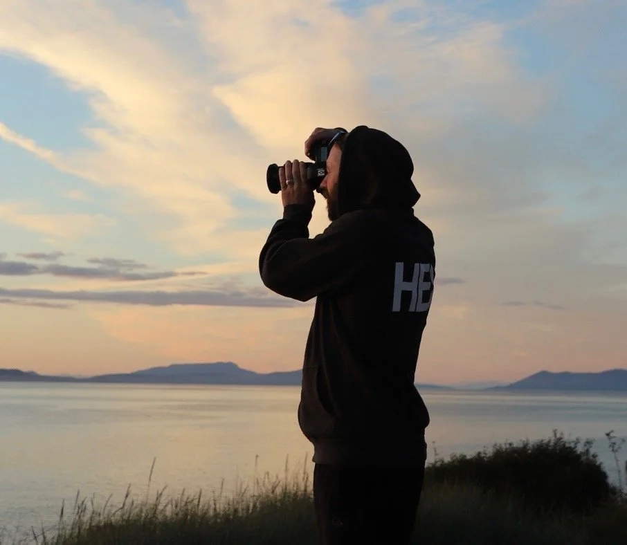Person in black hoodie taking a photo with a camera near a body of water during sunset.