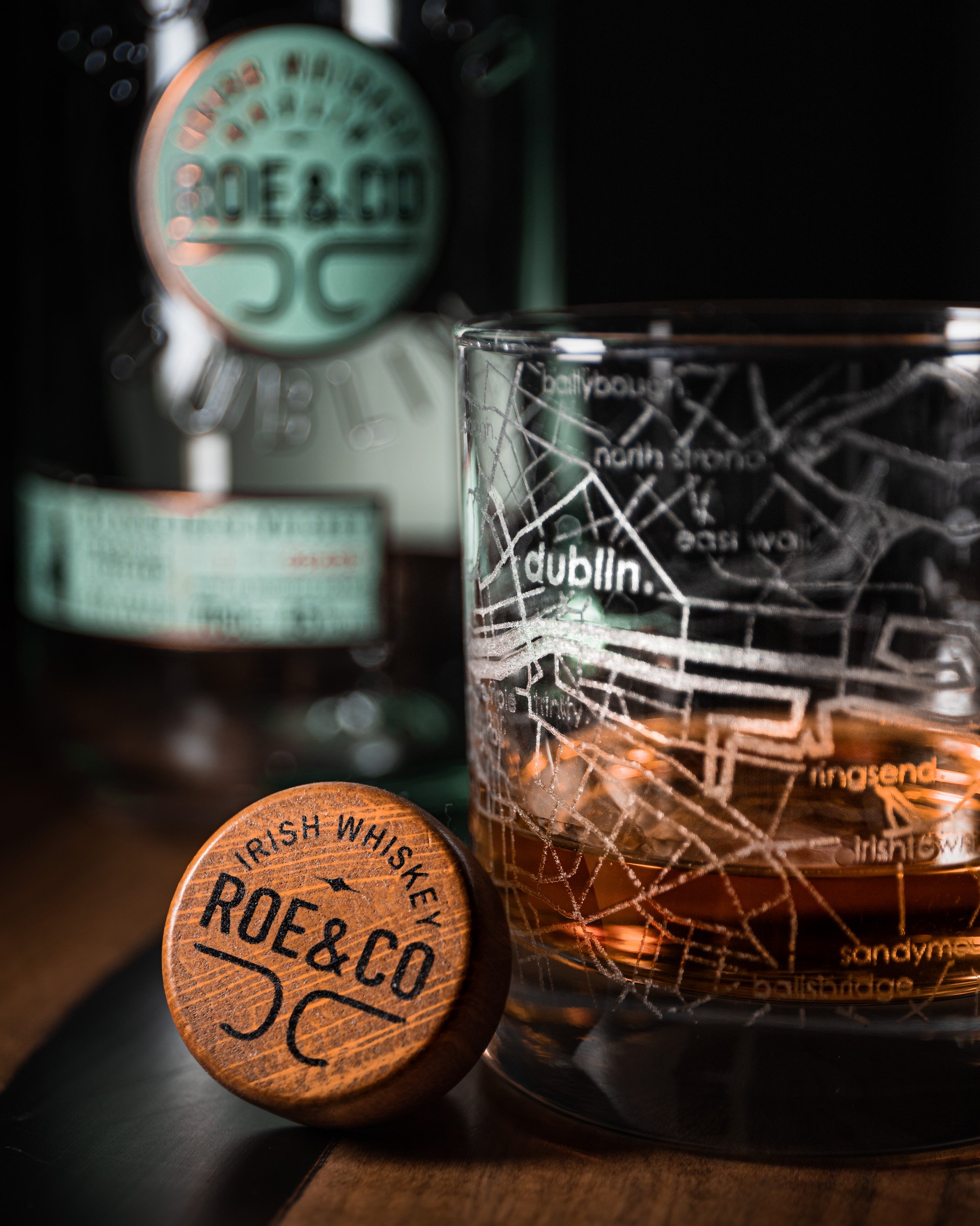 Close-up of a glass with a map design and the words 'dublin' and 'city' printed on it, along with a wooden coaster that says 'Irish Whiskey Roe & Co' in black text, and a bottle in the background labeled 'Jameson Irish Whiskey'.