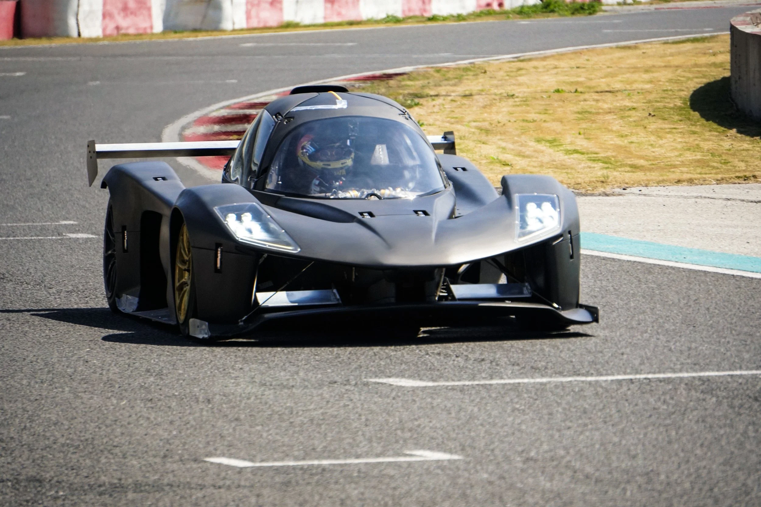 A sleek black racing car with a transparent canopy on a race track, driven by a helmeted driver visible through the canopy. The car has a large rear wing and yellow wheels, and is approaching a corner with red and white curbing.