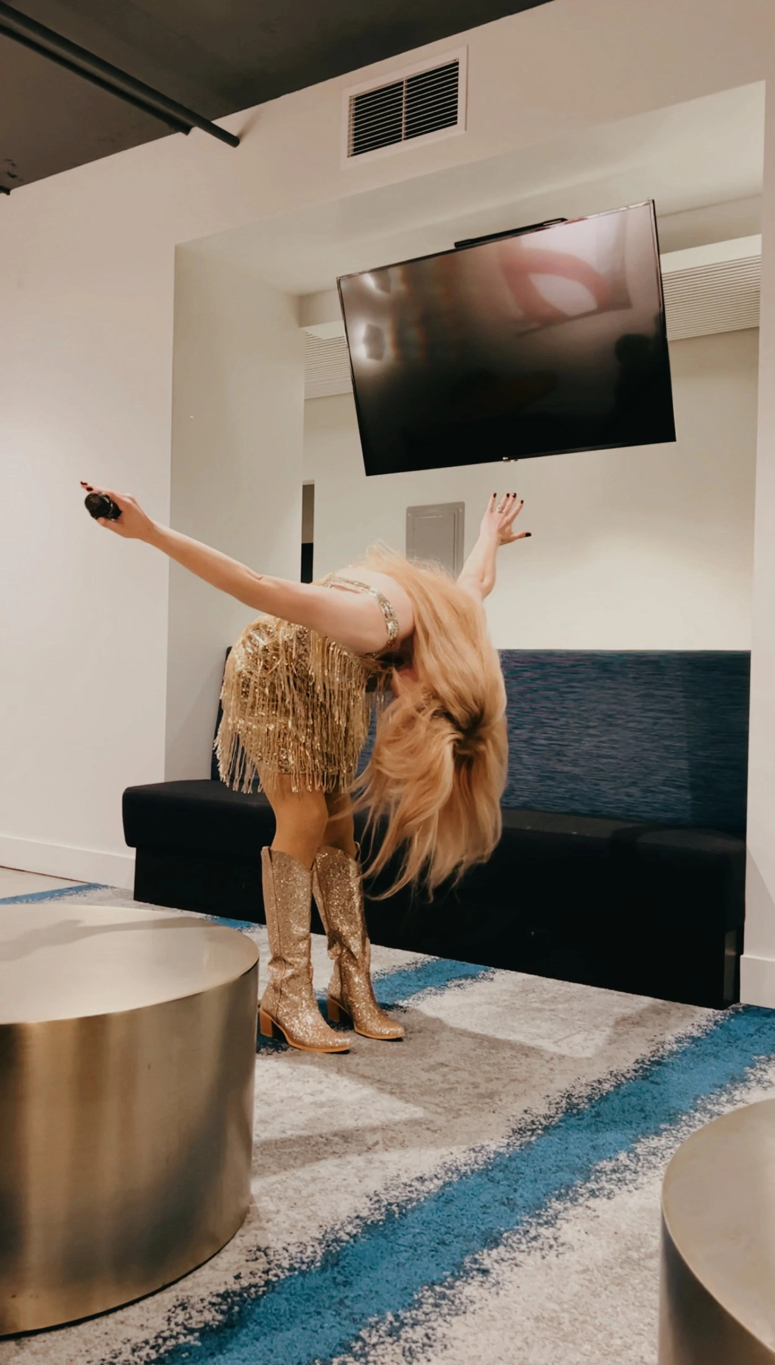 A person dressed in a gold sequined dress and matching gold glitter boots is bent over, with long blonde hair covering their face, in a modern indoor setting with a TV mounted on the wall and a black couch.