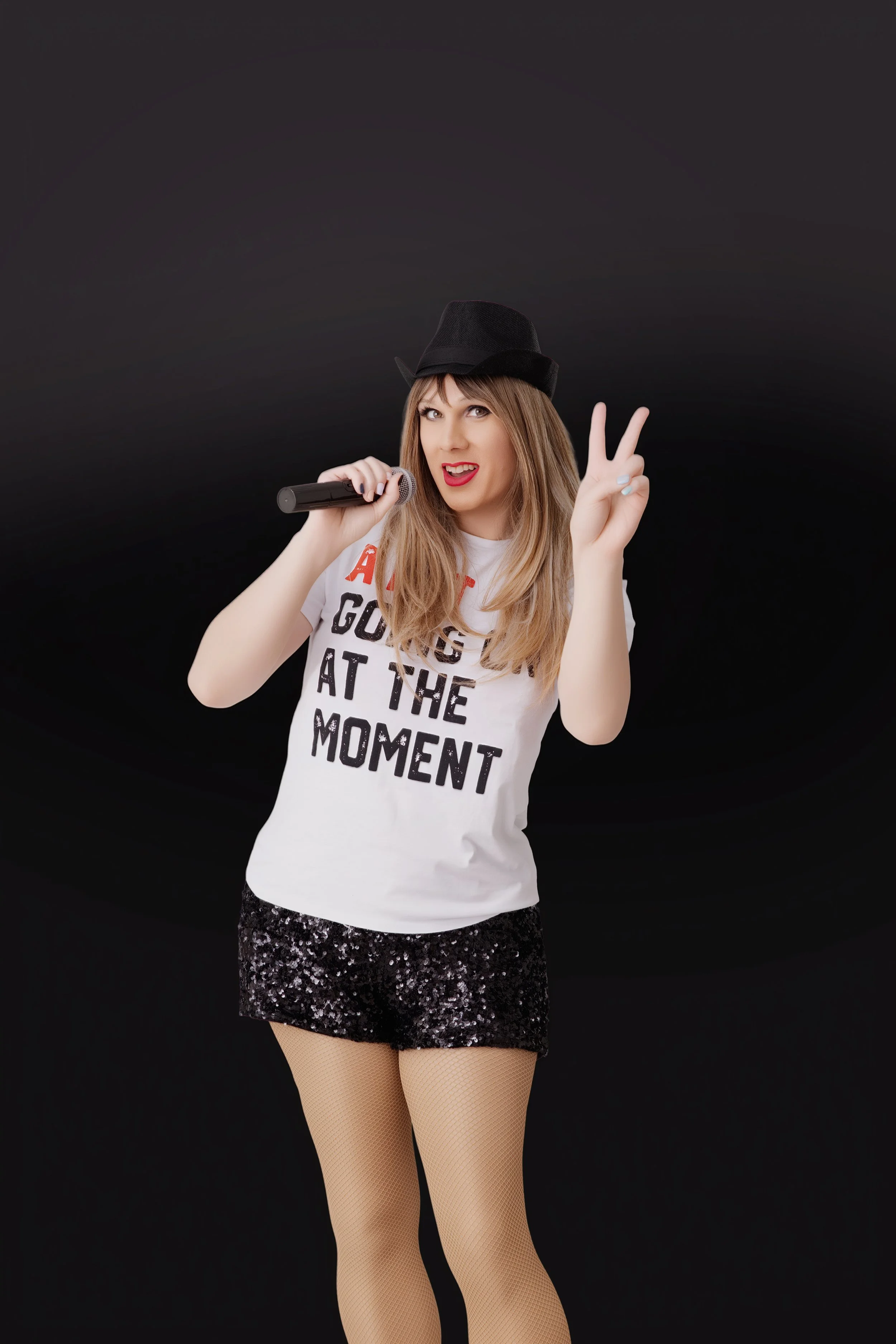 A woman holding a microphone and flashing a peace sign with her right hand. She is wearing a black hat, a white T-shirt with a slogan, a sequined black skirt, and fishnet tights, standing against a black background.