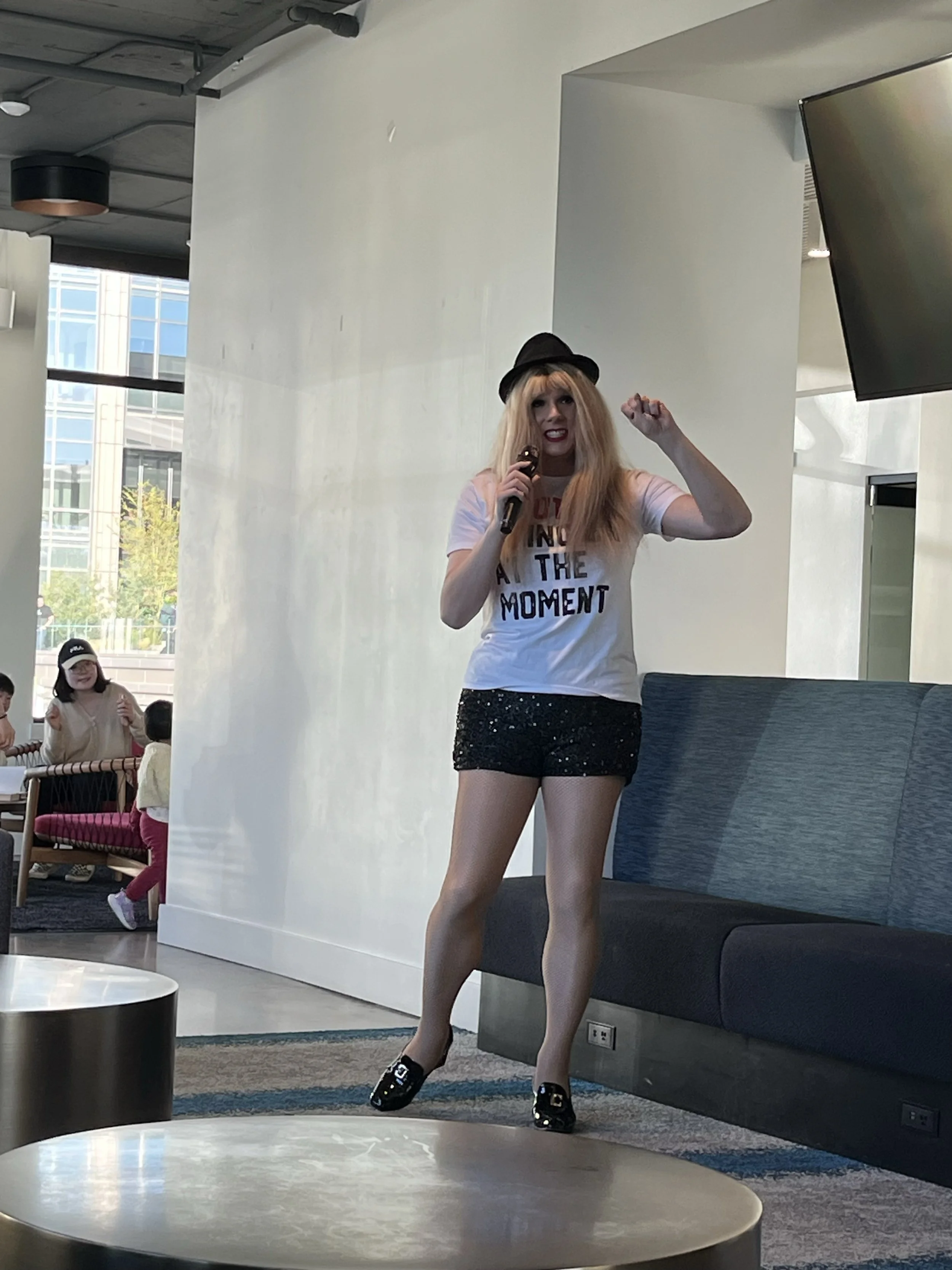 A woman with long blonde hair, wearing a black hat, a white T-shirt with bold text, and black shorts, is holding a microphone and performing or speaking in a modern indoor space with several people in the background.