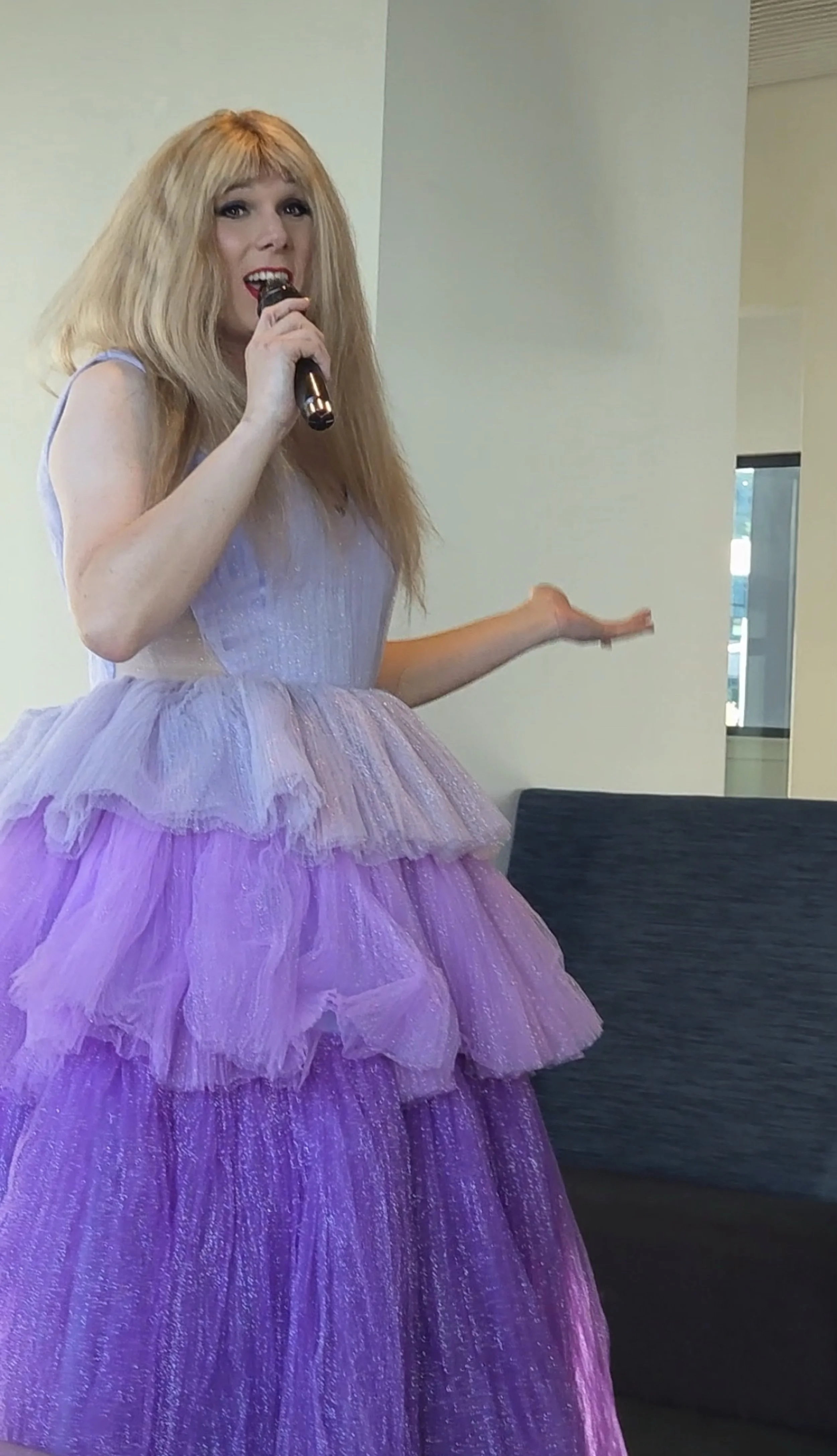Woman with long blond hair wearing a strapless purple, layered, tutu-style dress, holding a microphone, standing indoors near a wall and a window.