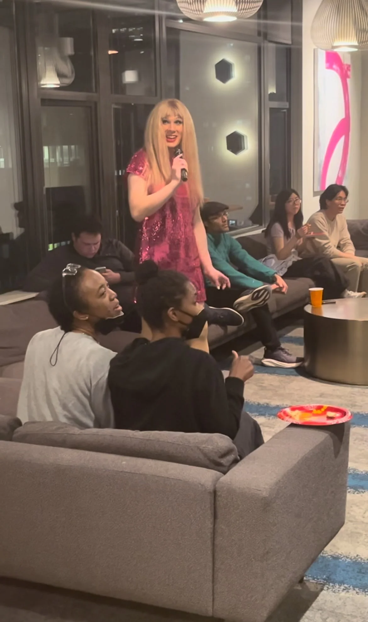 A person with long blonde hair and a pink sequin dress is standing and speaking into a microphone in a living room setting. Several people are sitting on couches, some with masks, and watching her. Modern decor includes large windows, hanging lights,