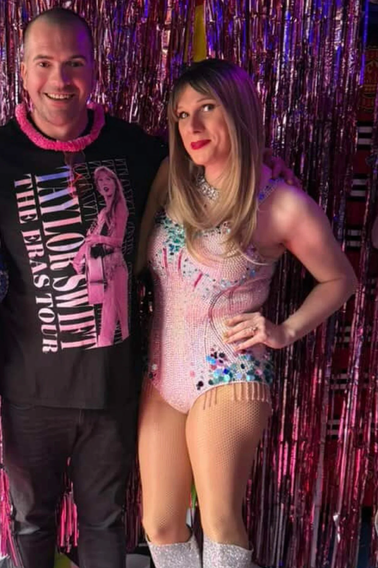 A woman wearing a colorful, sparkly leotard with a fishnet stocking and glittery knee-high socks standing next to a man in a black t-shirt with a pink necklace and graphic print, in front of shiny, metallic pink and purple decorations.