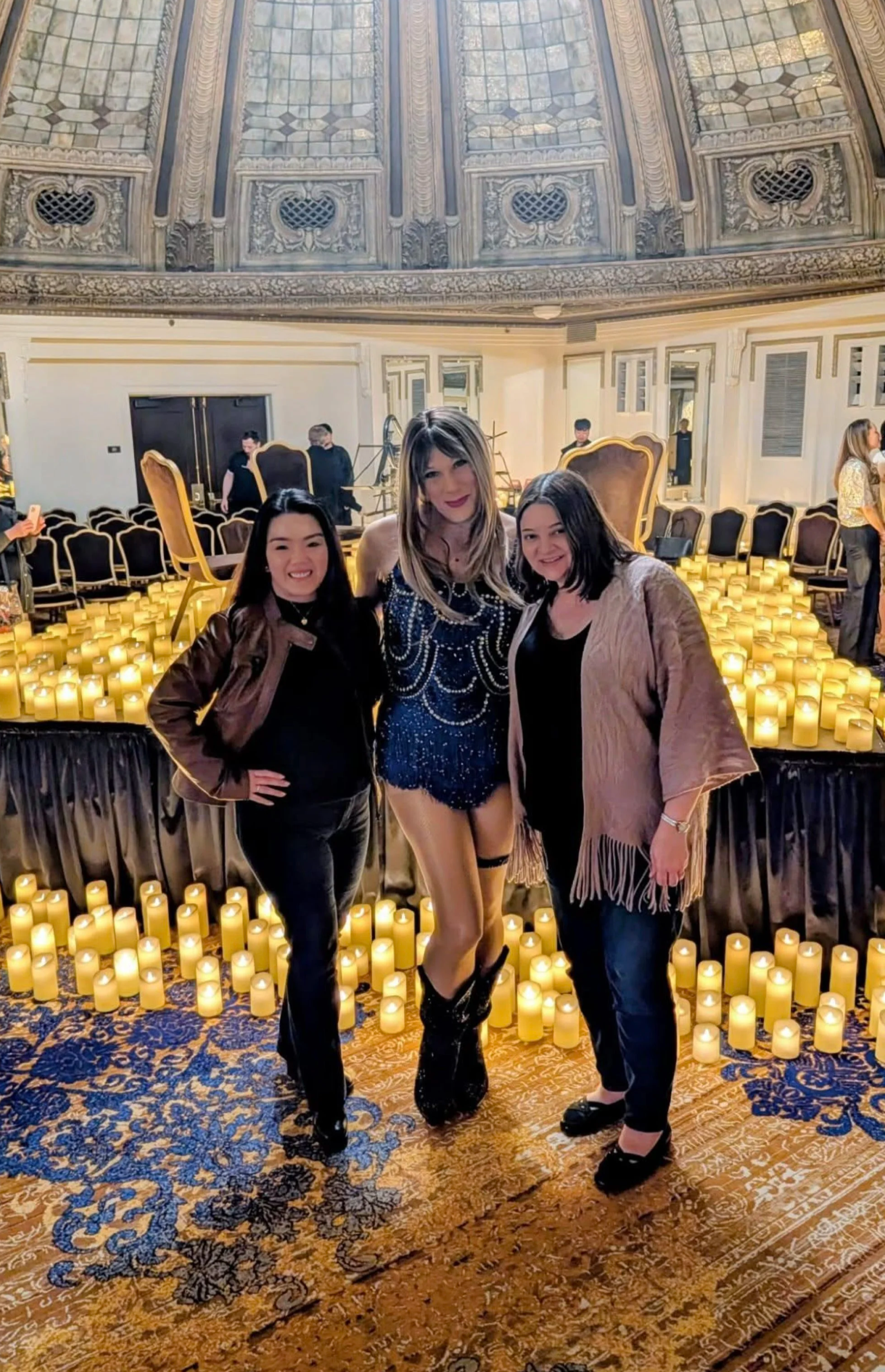 Three women standing together in front of a background decorated with candles and ornate architectural details, posing for a photo at an indoor event.