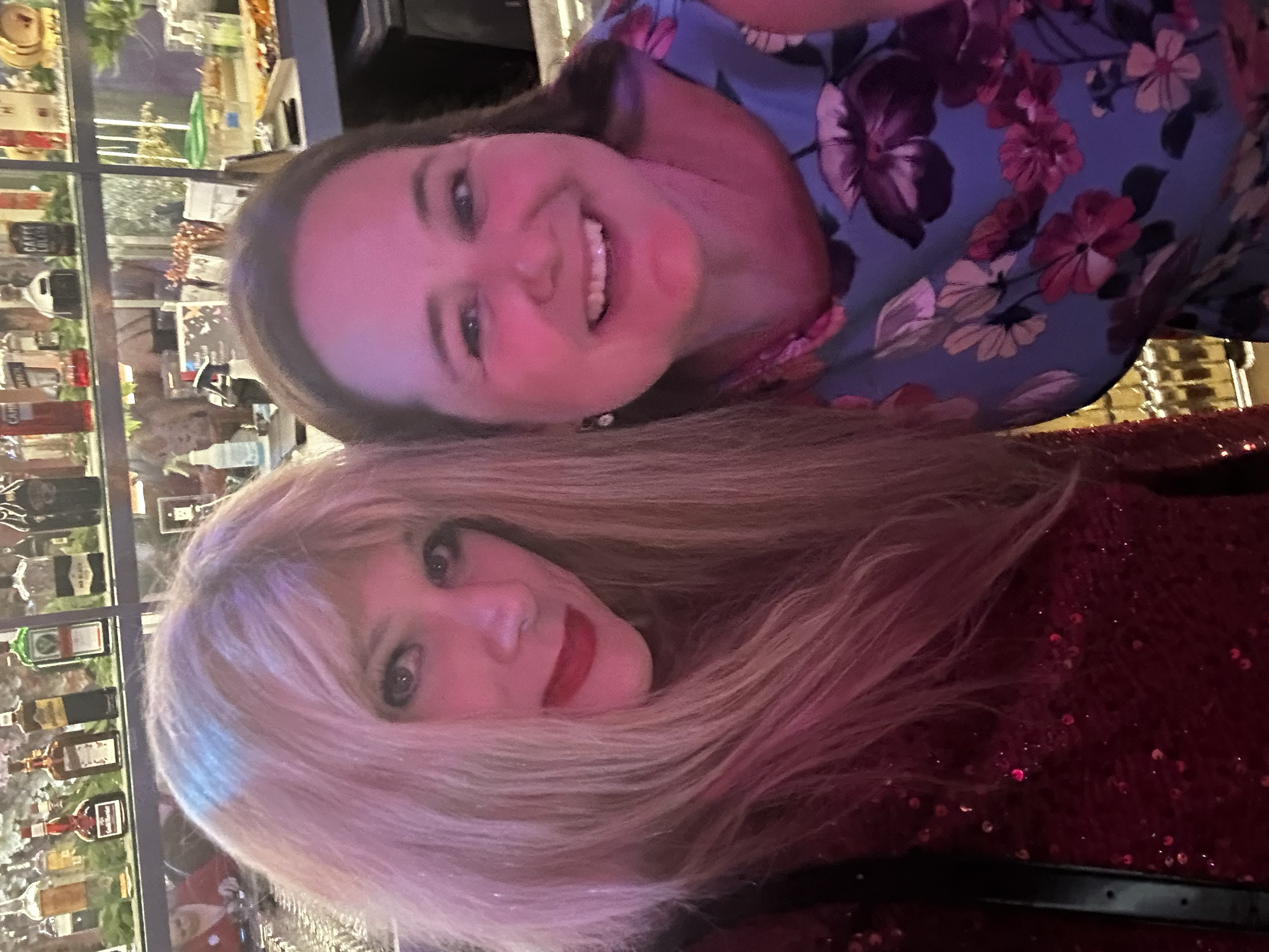 Two women with light hair smiling at a social gathering in a restaurant or bar, with a display of bottles and decorative jars in the background.