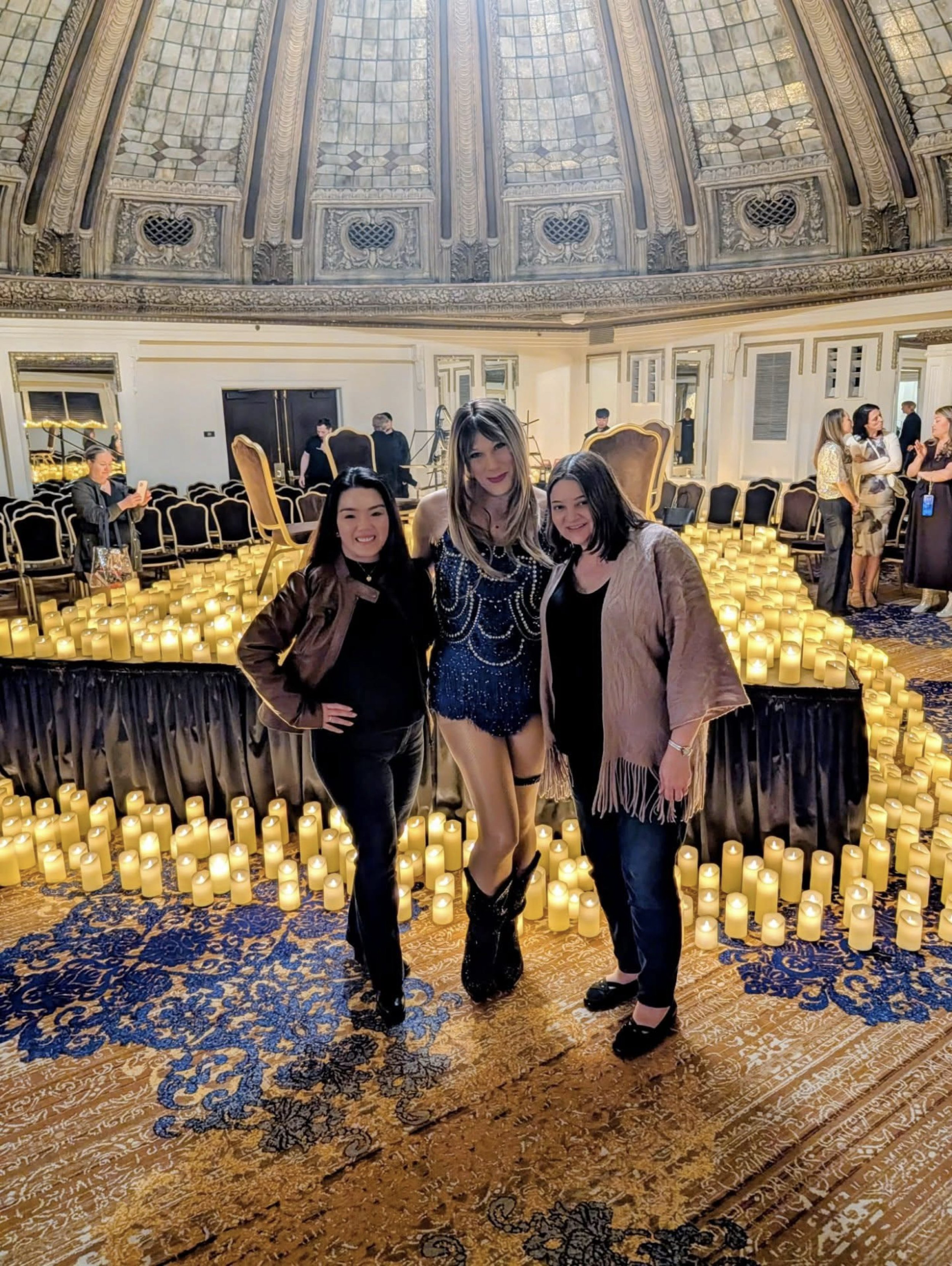 Three women stand together in front of a candle display inside an ornate hall with a domed ceiling and a patterned carpet.