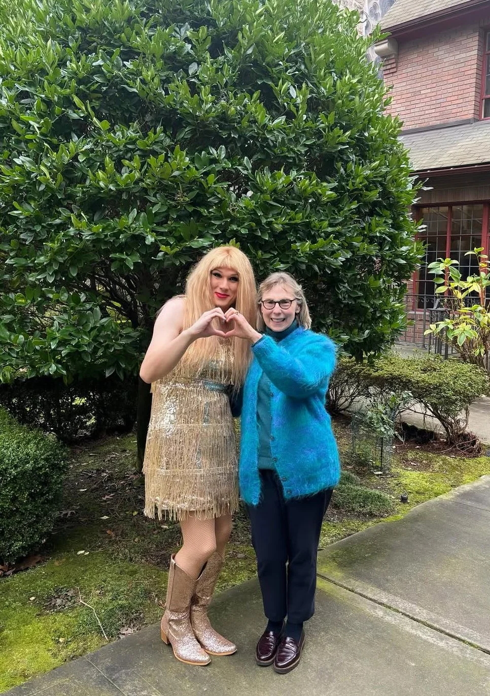 Two women standing in front of a large green bush, making a heart shape with their hands.
