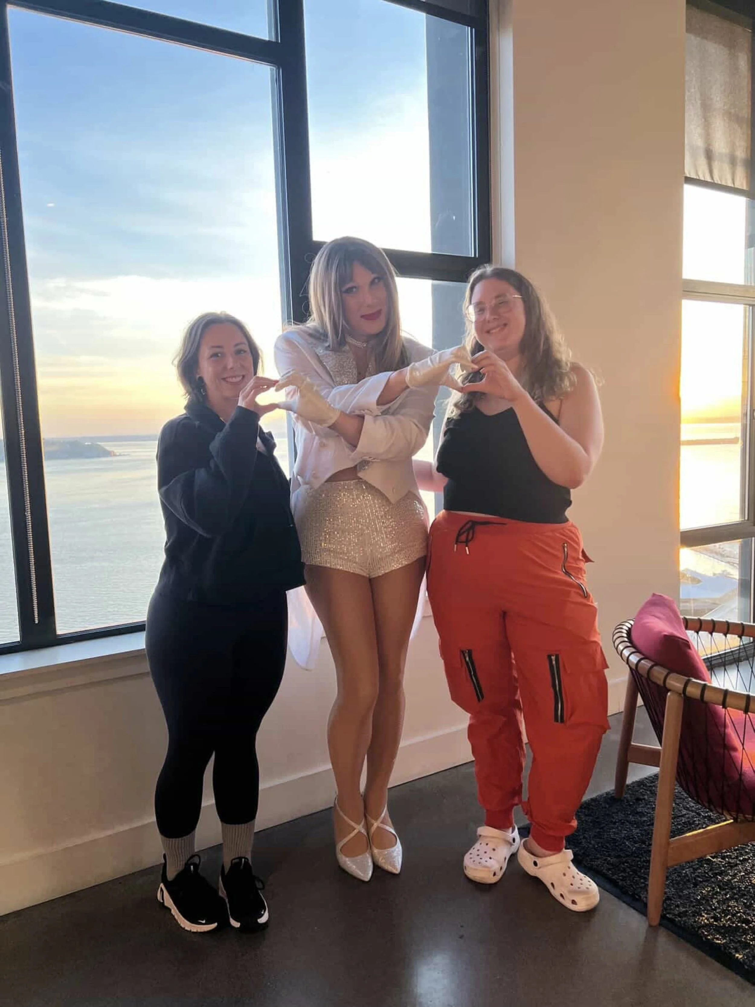 Three women standing indoors by a large window with a scenic view of water and sky, forming a heart shape with their hands. The woman in the middle is dressed in a shiny, fashionable outfit, and the other two women are dressed casually.