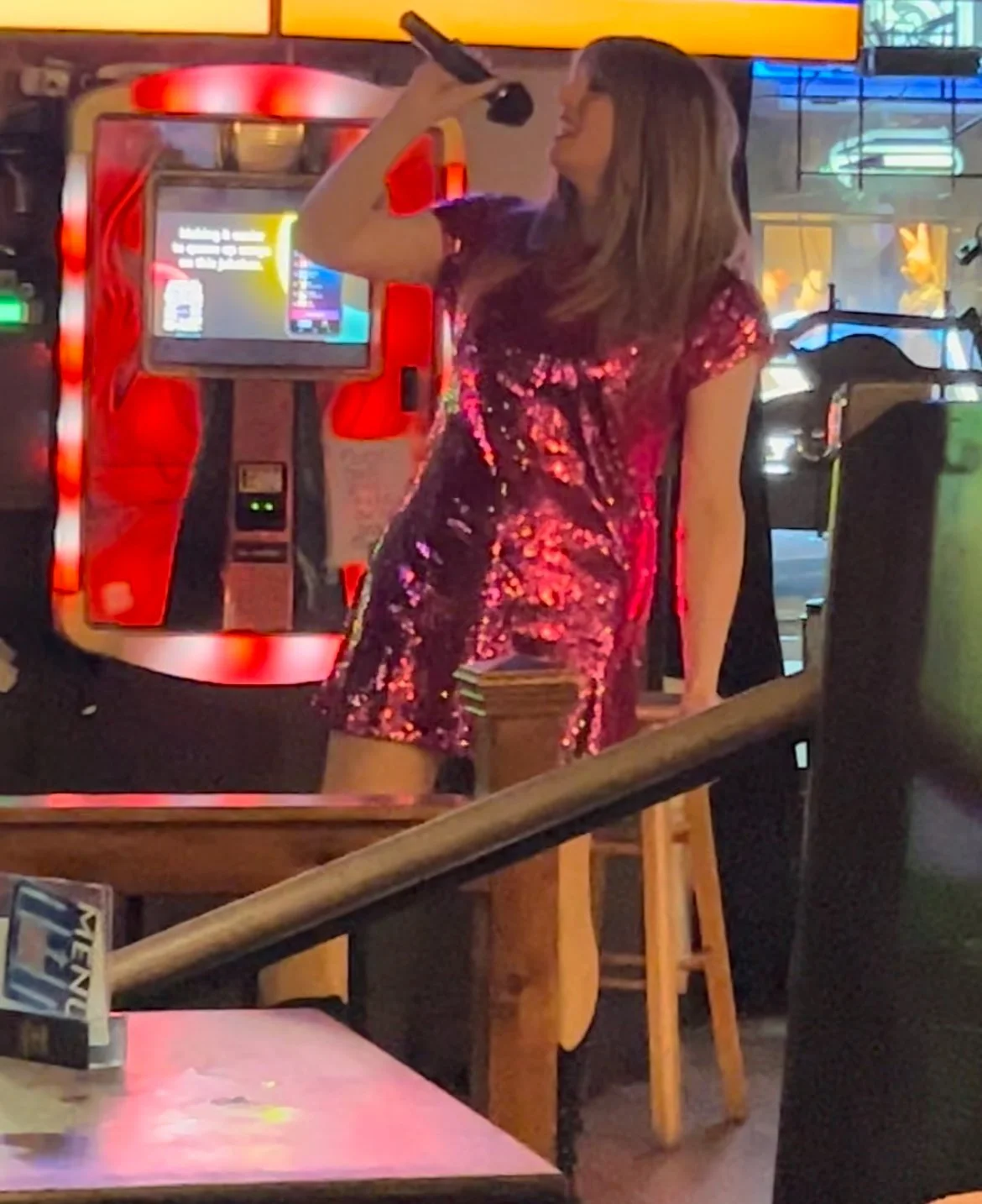 A woman with long hair wearing a shiny, red and black dress, singing into a microphone at an indoor entertainment venue with neon lights and an ATM in the background.