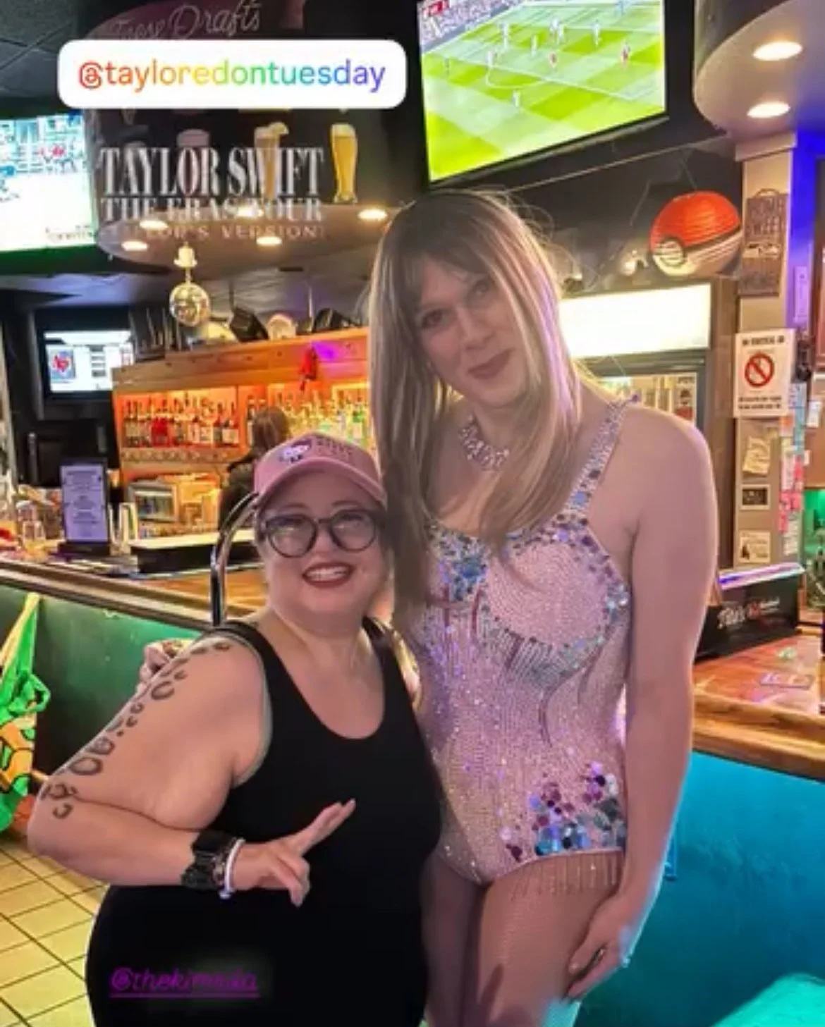 Two women standing inside a sports bar, smiling at the camera. One woman is wearing a black top and glasses with a pink cap, and the other is wearing a sparkling dress. There are multiple TV screens showing sports, a bar counter with drinks, and deco